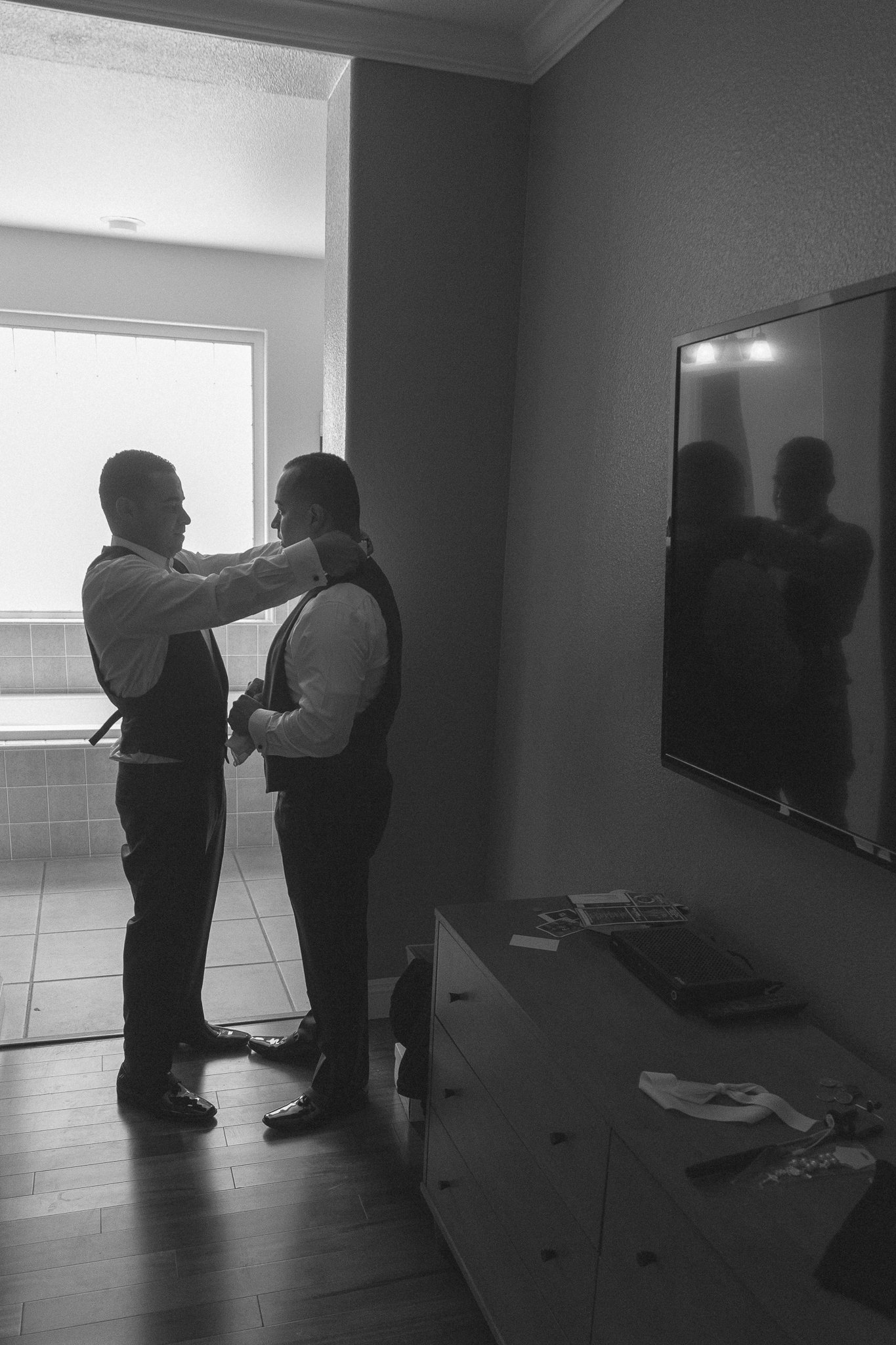Two men in formal attire, one adjusting the bow tie of the other, standing in a room with a TV and dresser, preparing for an event.