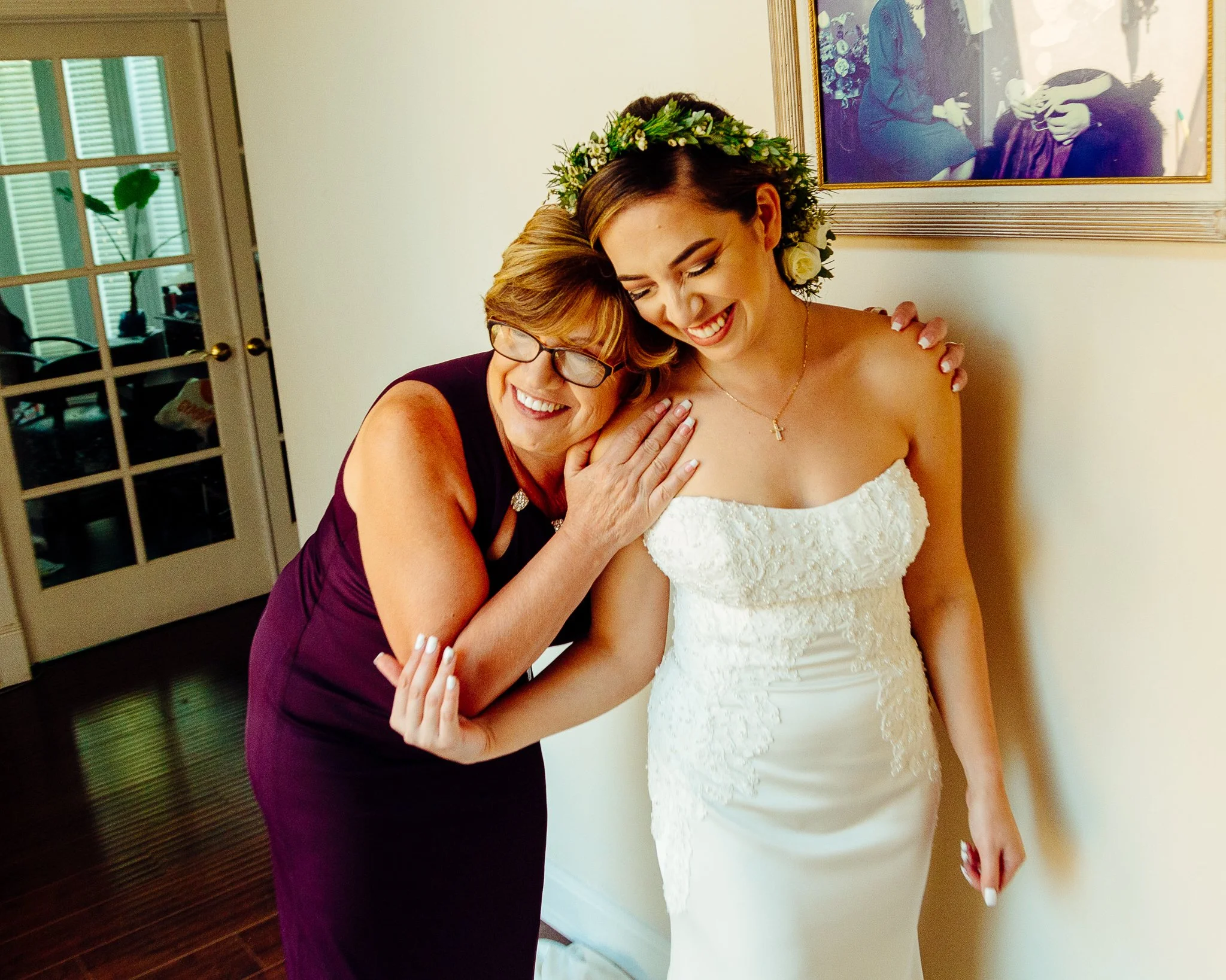 A smiling woman in a white wedding dress hugging an older woman in a purple dress, who is also smiling; the bride wears a floral crown and has her eyes closed, leaning into the hug.
