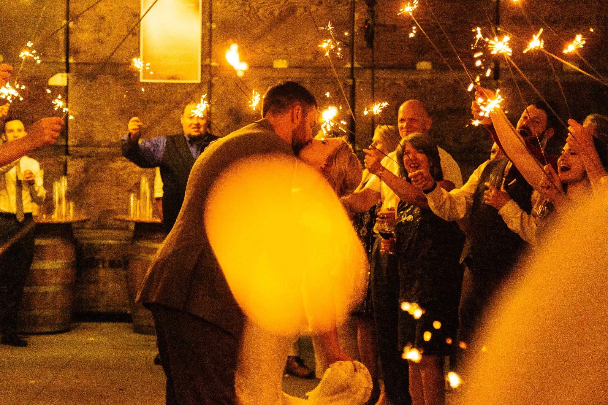 A couple in wedding attire sharing a kiss amidst a celebration with sparklers held by friends and family. The background shows a rustic brick wall and barrels, creating a festive atmosphere.
