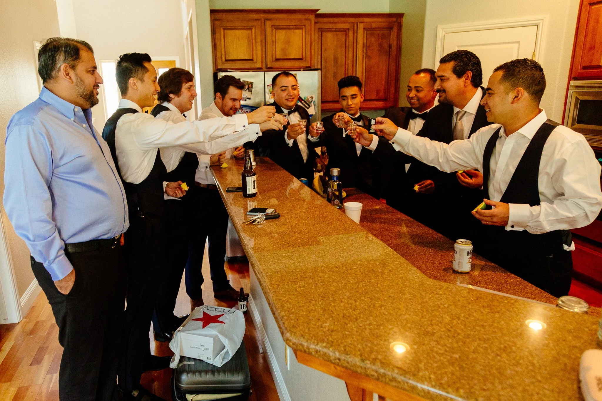 A group of men dressed in formal attire, toasting with drinks at a bar in a home kitchen.