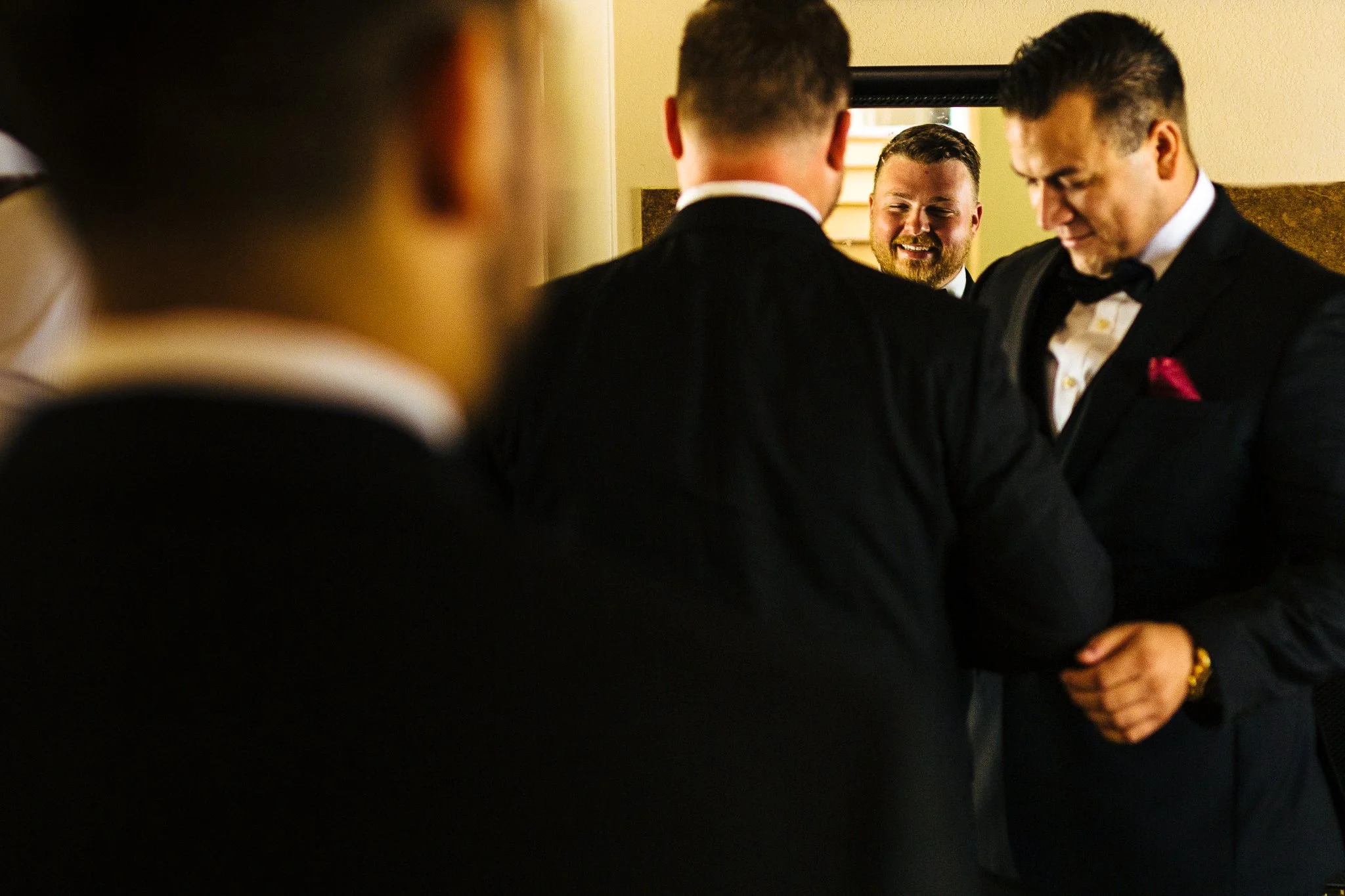 A group of men in tuxedos, some with bow ties, gathered together in a room, smiling and engaging with each other, perhaps during a wedding or formal event.