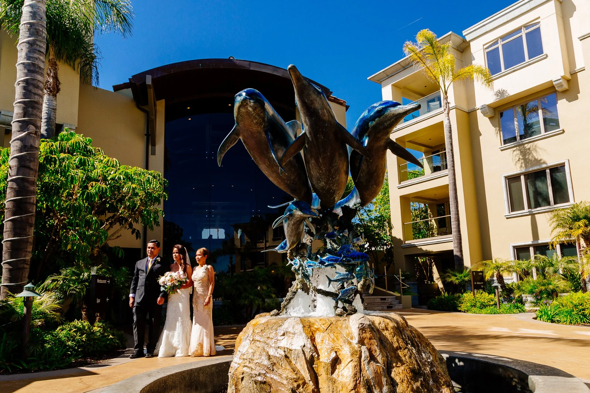 A wedding couple standing near a fountain with dolphin sculptures outside a modern building, surrounded by tropical plants and palm trees.