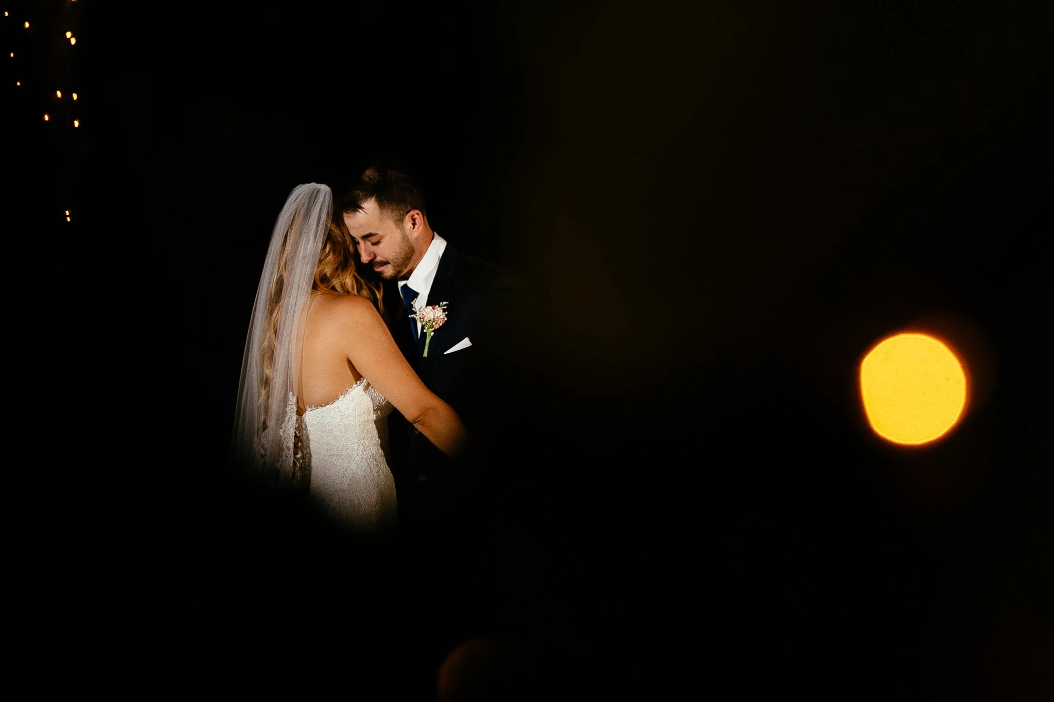 A bride and groom sharing a moment at their wedding, with the bride wearing a white lace dress and veil, and the groom in a dark suit with a boutonniere, against a dark background with a blurred orange light.