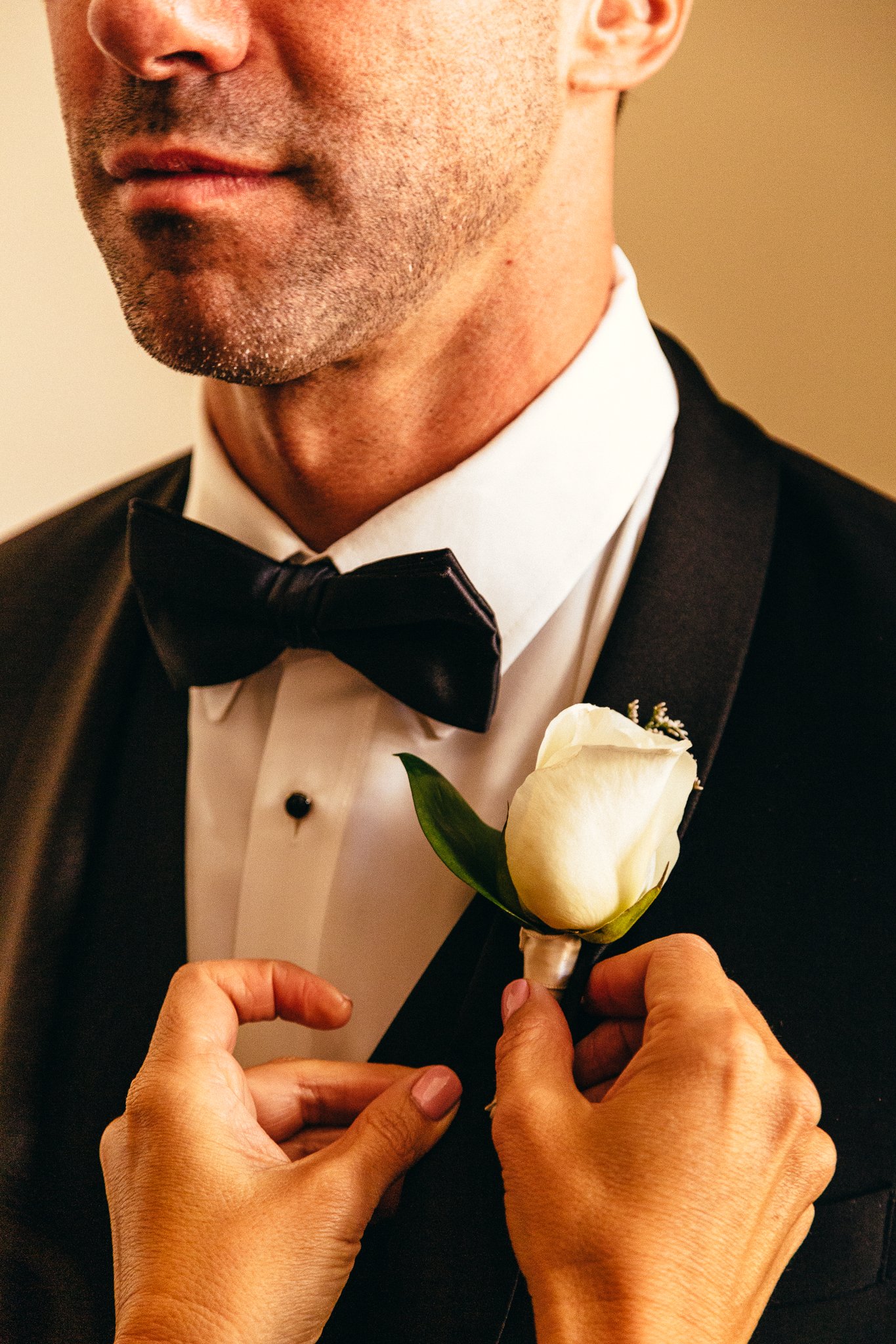 A man in a tuxedo with a bow tie, adjusting a white boutonniere on his lapel, likely preparing for a formal event or wedding.