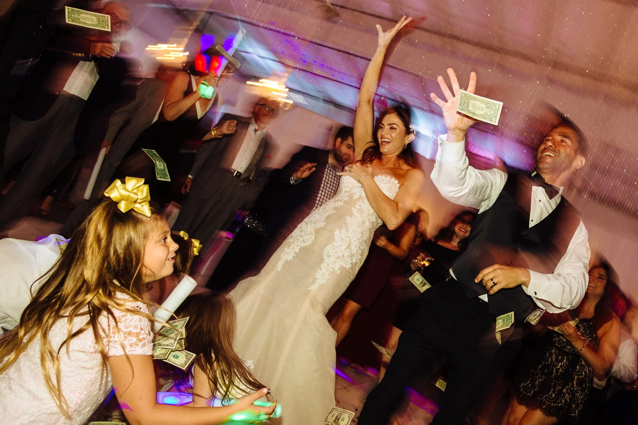 Wedding celebration with bride and groom dancing, surrounded by guests, with dollar bills falling, and children enjoying the party.