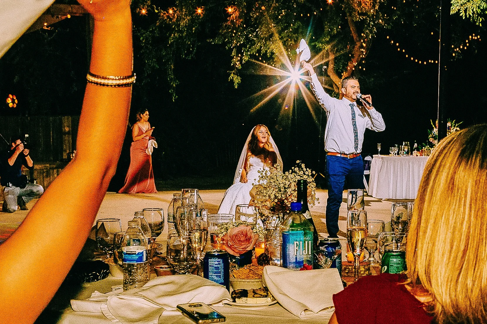 A wedding celebration at night outdoors, with a bride in a white dress and veil sitting among flowers, a man in a shirt and tie giving a speech with a microphone, and a woman in a pink dress standing nearby. The table in the foreground has glasses, b