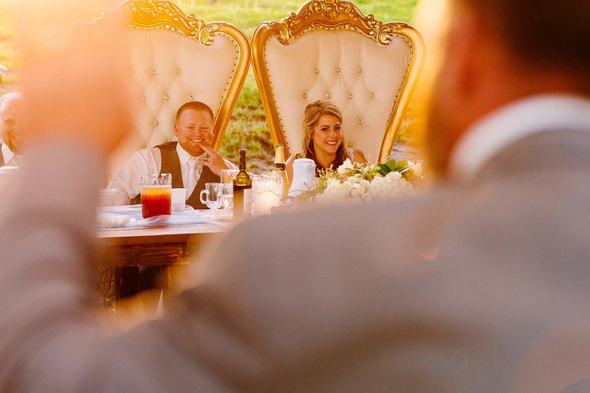 A bride and groom sitting at a head table during their wedding reception. The bride is smiling and the groom is smiling, surrounded by people. The scene is outdoors with a decorated, gold-accented backdrop and floral arrangements.