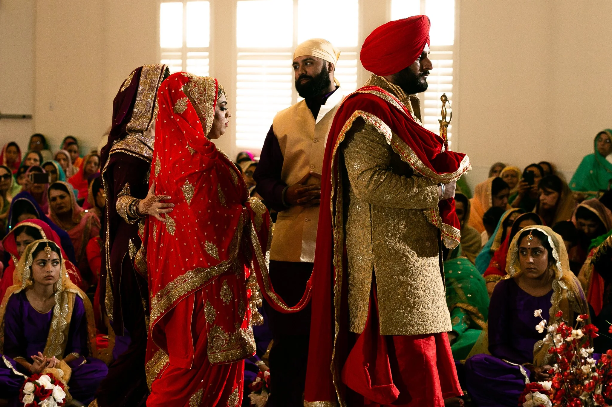 Indian wedding ceremony with the bride and groom dressed in traditional attire, surrounded by seated guests wearing colorful sarees and salwar kameez in a decorated indoor setting.