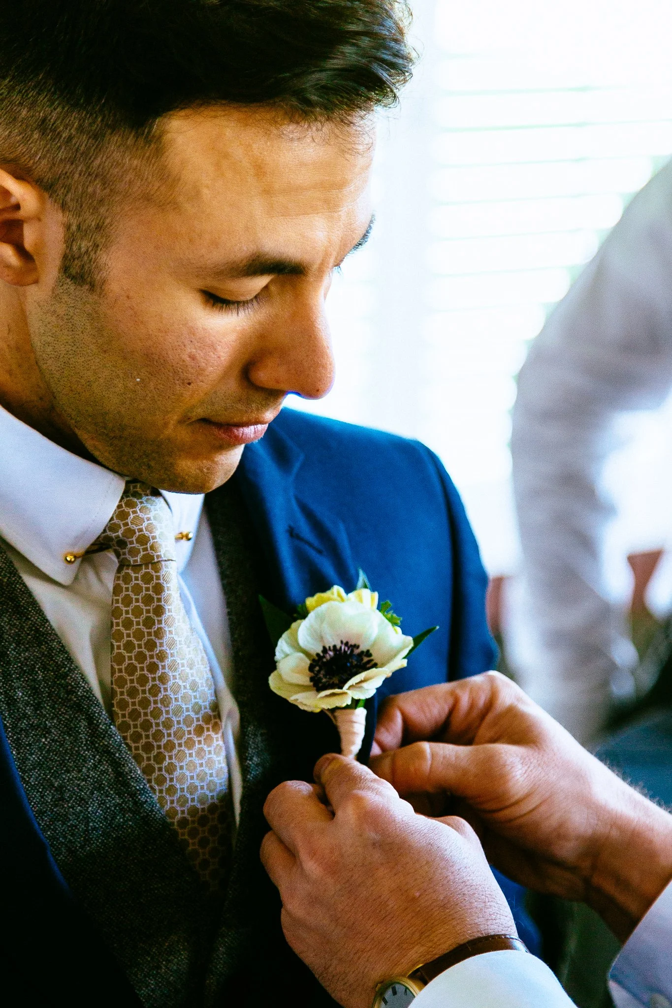 A man in a blue suit is getting a white flower pinned to his jacket by someone with a gold wristwatch, with a bright window in the background.