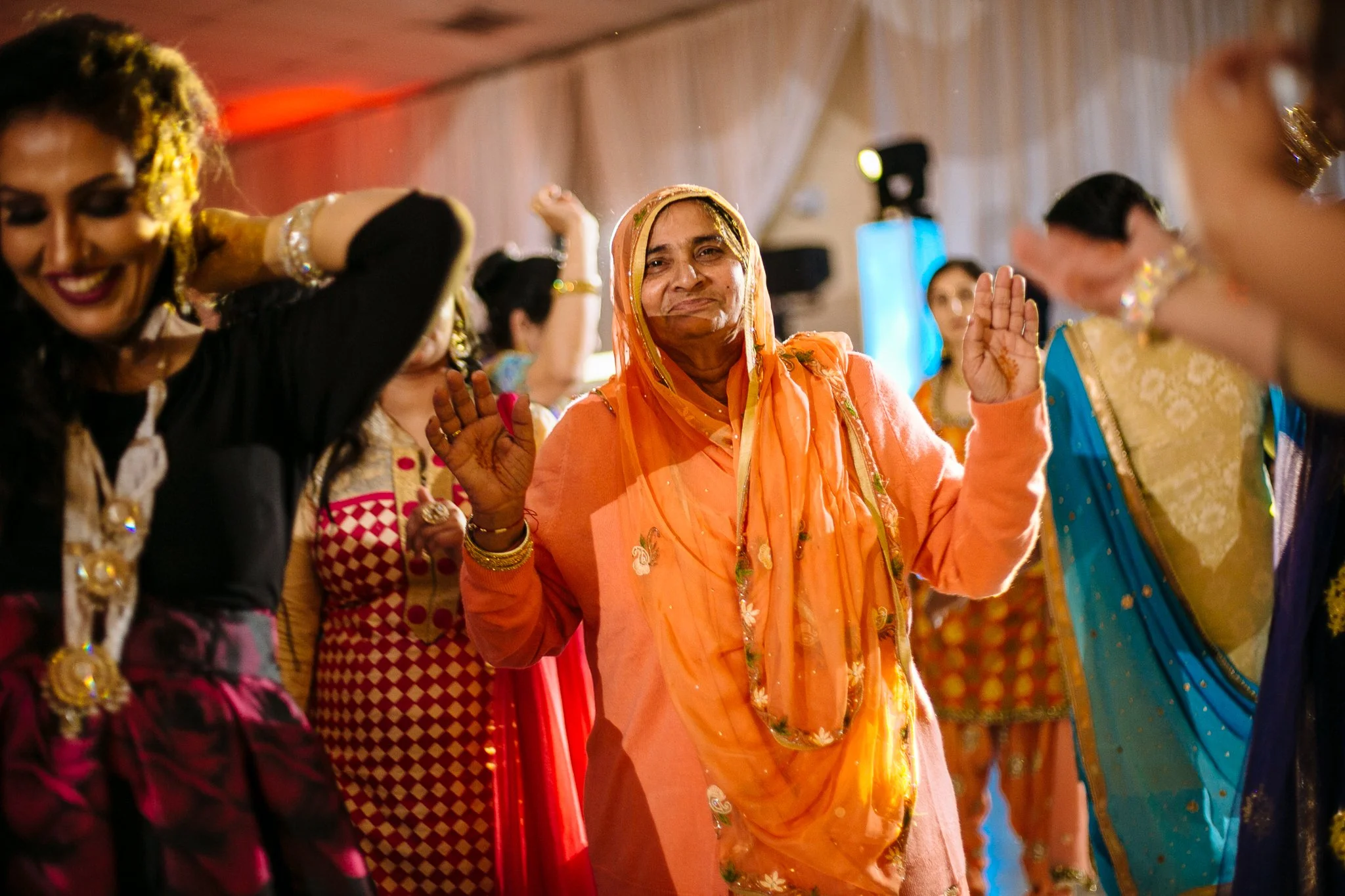 Women dressed in colorful traditional Indian clothing dancing at a celebration, with the woman in the center wearing an orange saree and smiling.