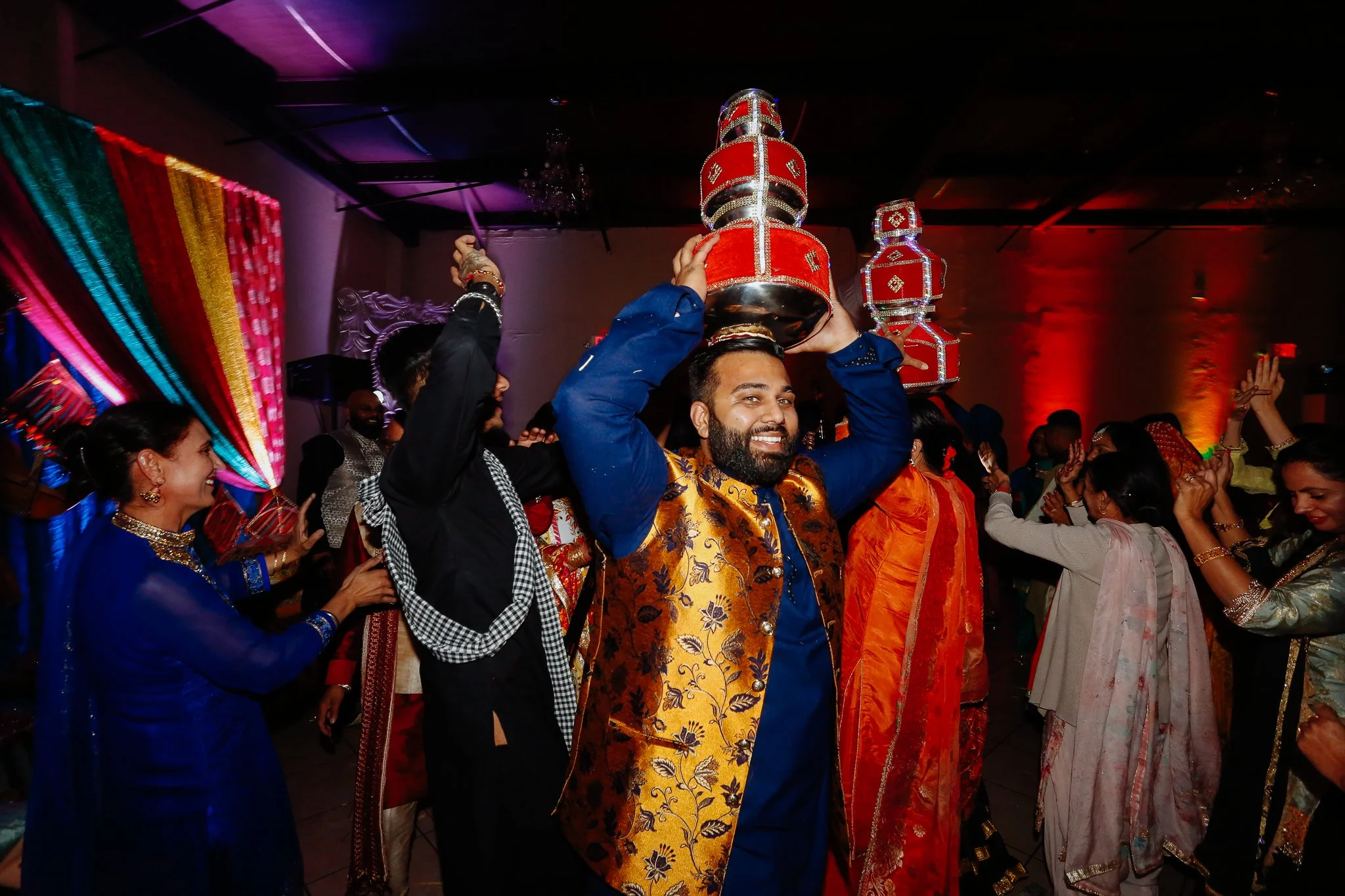 A man smiling and wearing a colorful traditional outfit, balancing decorated containers on his head at an Indian celebration with others dancing and celebrating around him.