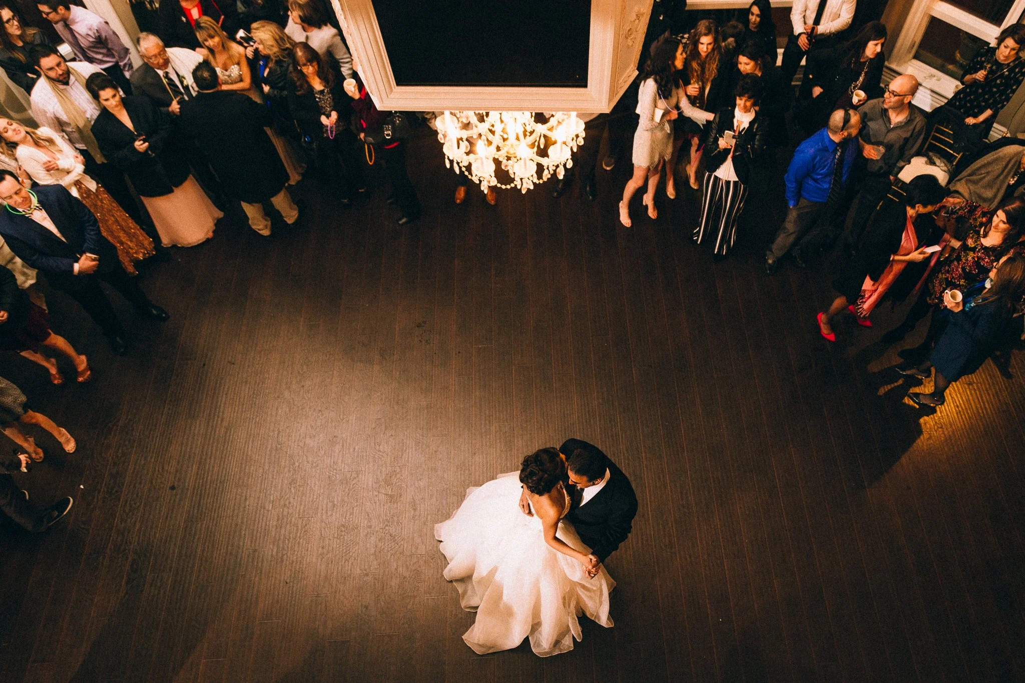 A bride and groom dancing in the center of a dark wood floor at their wedding reception, surrounded by guests standing along the edges of the dance floor watching them.