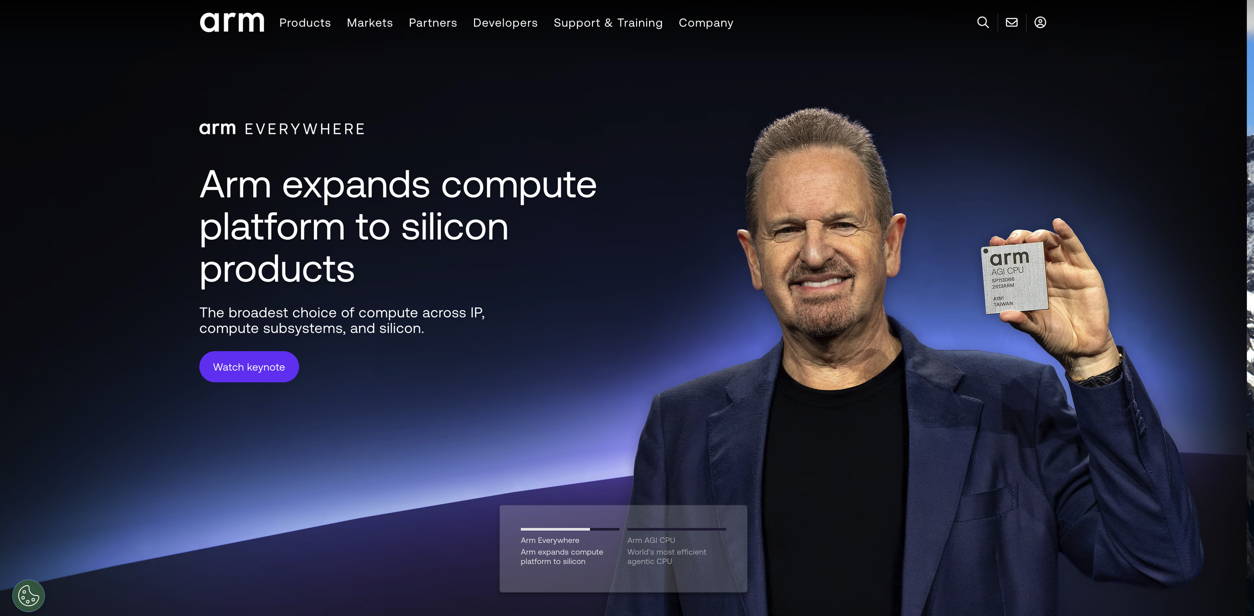 A man in a dark blazer is holding a small square computer chip with the brand name 'arm' printed on it. The background is dark, and there is a website header with navigation links at the top.