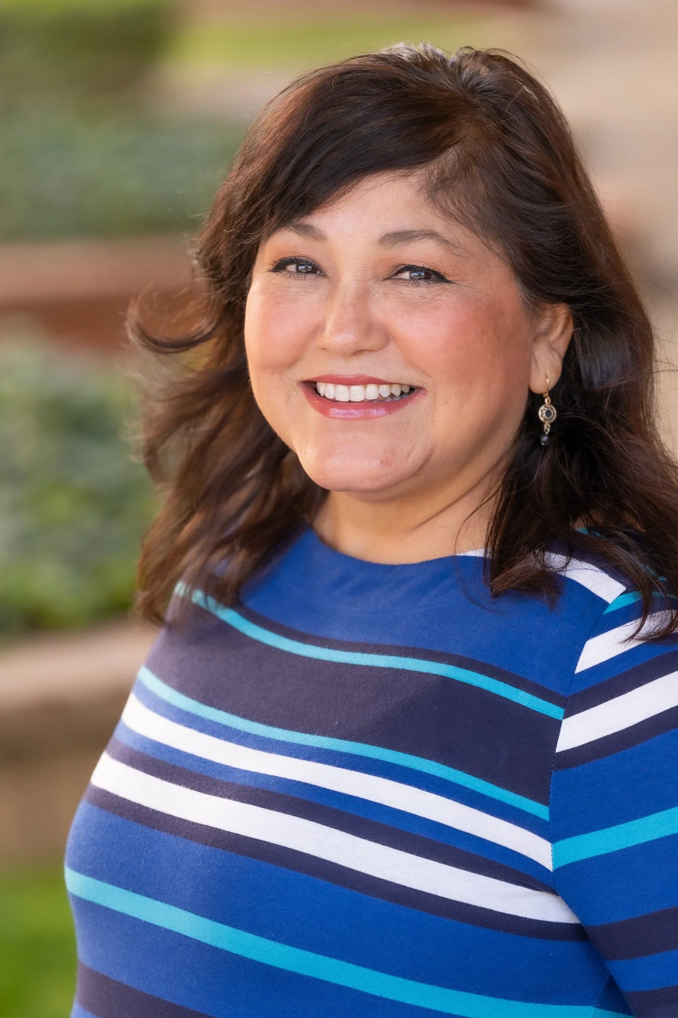 A woman with shoulder-length brown hair, smiling, wearing a blue striped top and earrings, outdoors with blurred greenery in the background.