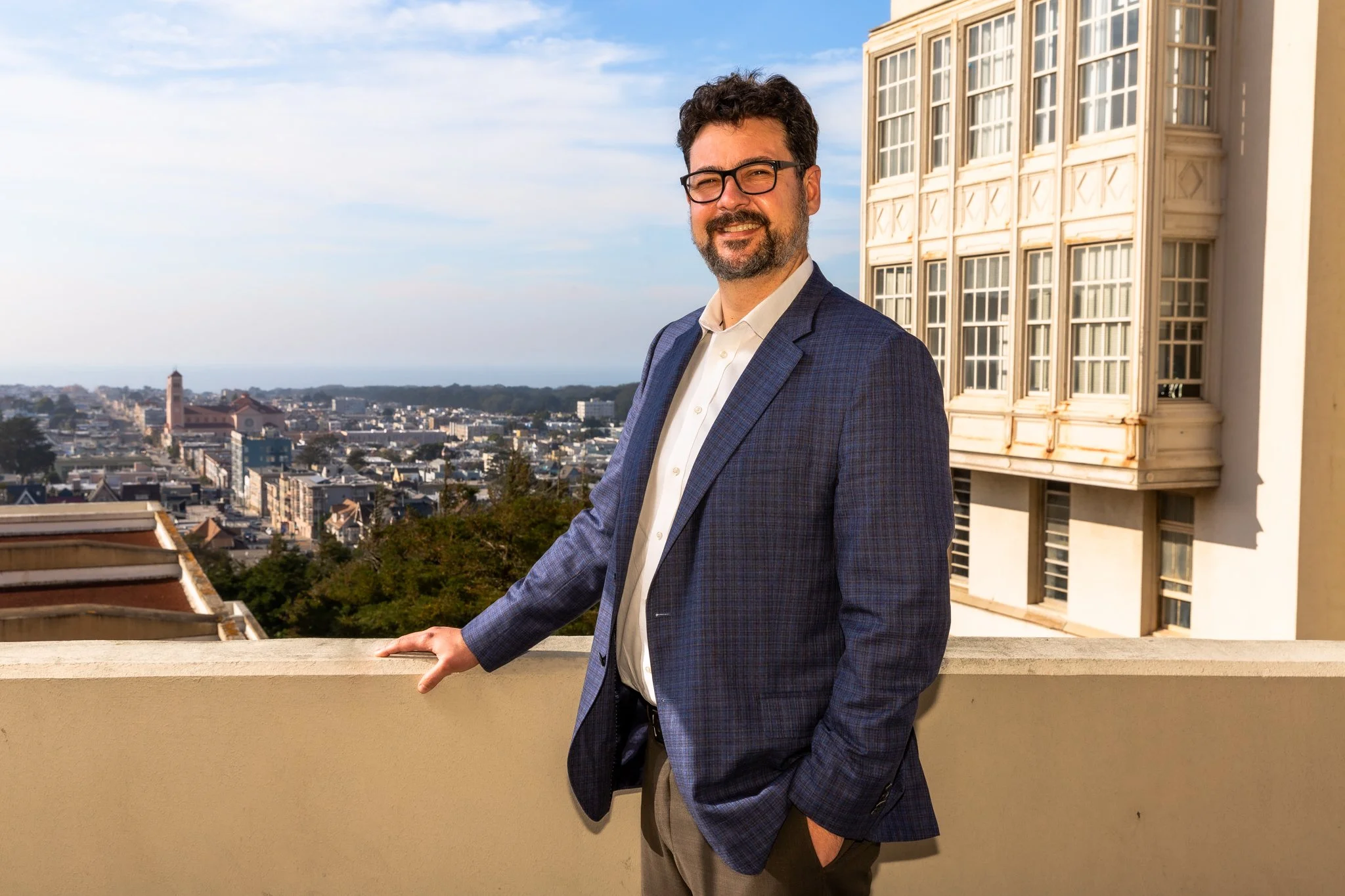 A man with dark hair, glasses, and a beard, wearing a blue plaid blazer and light-colored pants, stands on a balcony with his right hand resting on the railing. The background shows a city skyline with various buildings under a partly cloudy sky.