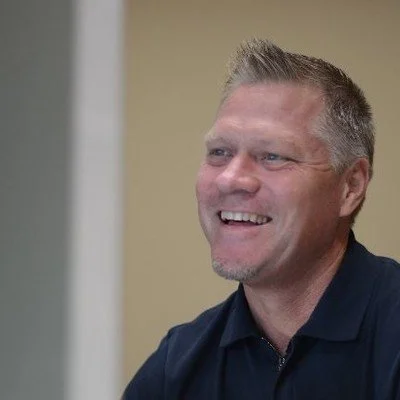 A smiling man with short gray hair wearing a navy blue collared shirt.