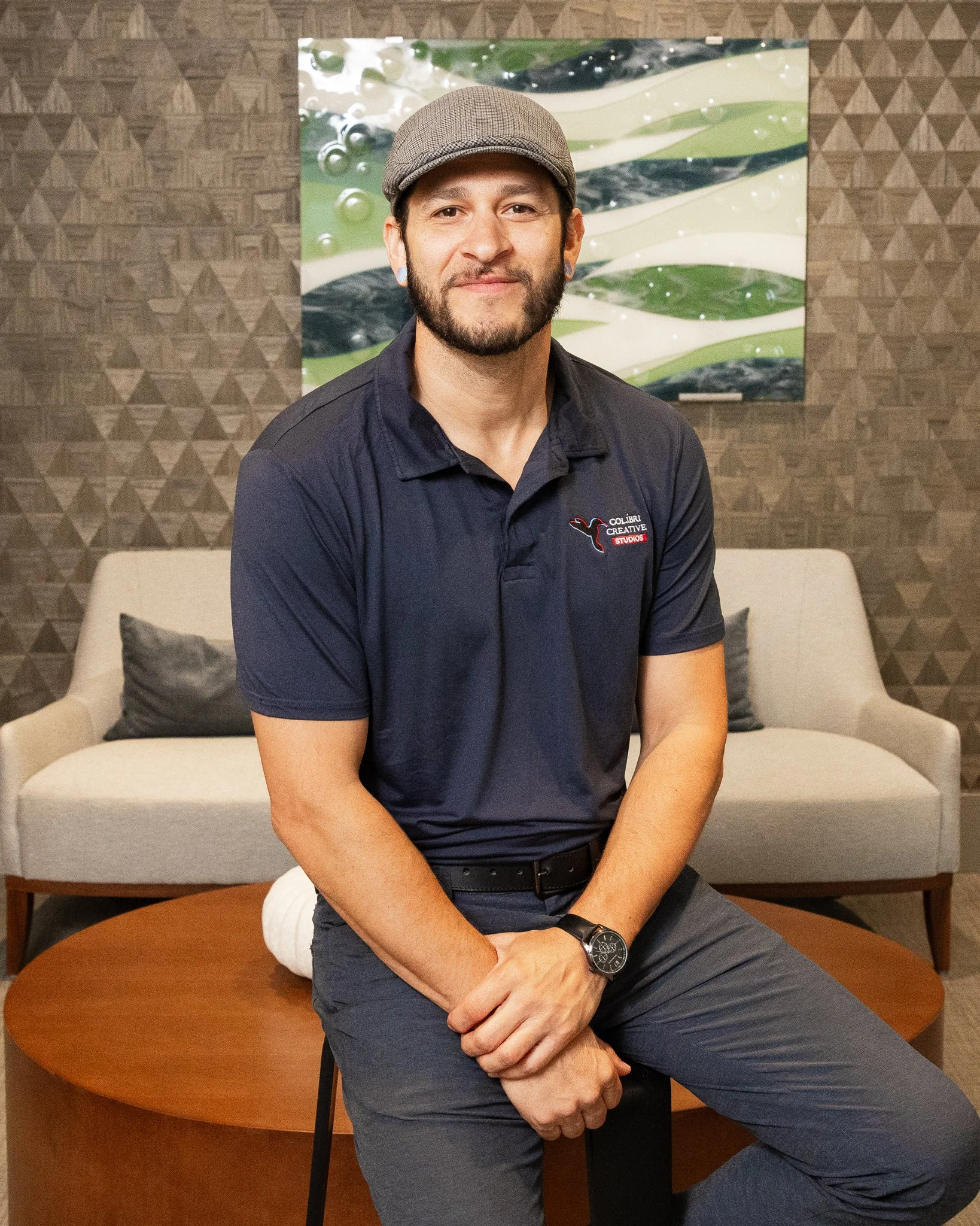 A man with a beard and mustache wearing a gray flat cap, navy polo shirt with a logo, gray pants, and a watch, sitting on a stool with a beige couch and a modern abstract painting in the background.