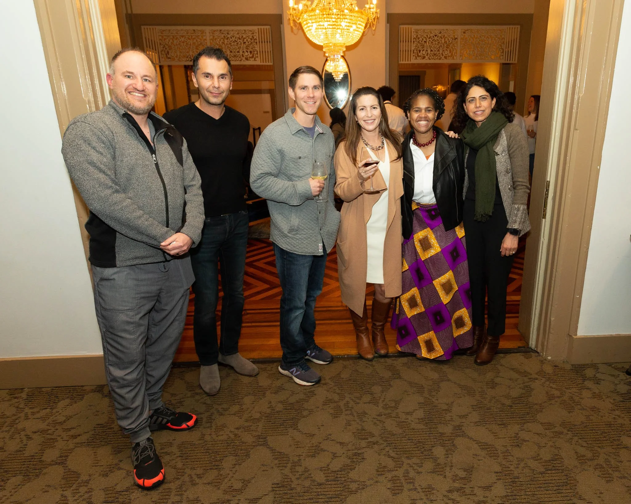 A group of seven people standing together at an indoor social event, with two women holding glasses of wine, smiling for the camera, in front of a chandelier and ornate background.