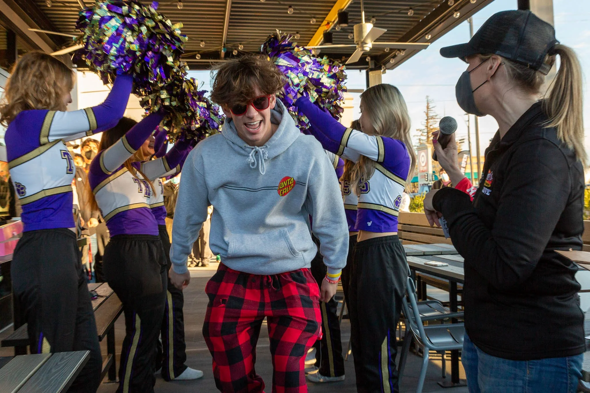 A young man with red sunglasses smiling and dancing as cheerleaders hold pom-poms above his head. A woman with a microphone wearing a face mask is talking to him. They are at an outdoor event with tables and a partial view of a chain link fence and a