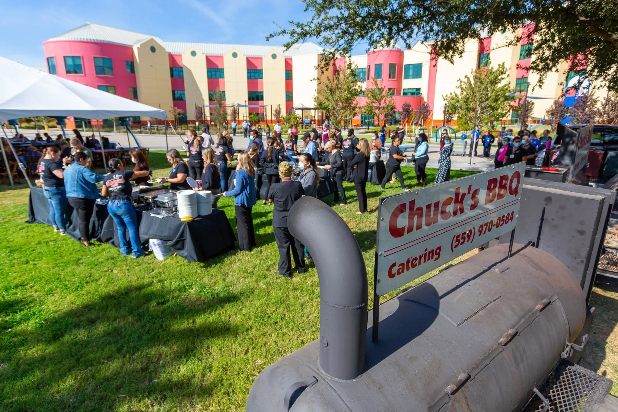 Outdoor gathering at a park with a food buffet run by Chuck's BBQ catering; large group of people lined up, some wearing masks, in front of a colorful modern building on a sunny day.