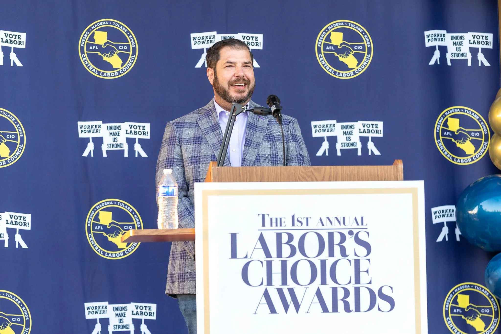 A man standing at a podium decorated with a sign that reads 'The 1st Annual Labor's Choice Awards,' speaking into a microphone. The background features banners with logos of the Central Labor Council, AFL, CIO, and supportive slogans such as 'Vote La