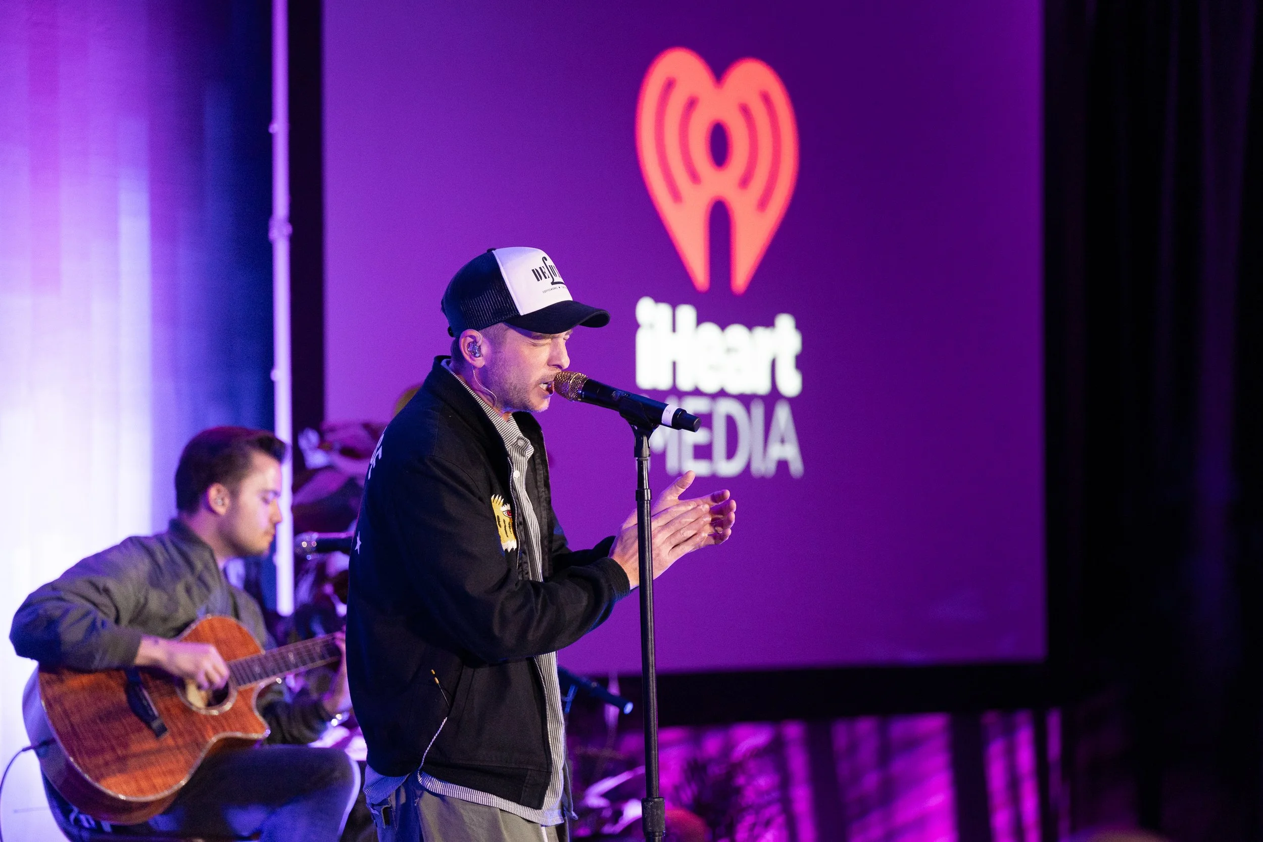 A male singer wearing a black cap and black jacket performing on stage with a microphone, a guitarist sitting beside him playing an acoustic guitar, and a large illuminated iHeartMedia logo projected on a screen in the background.