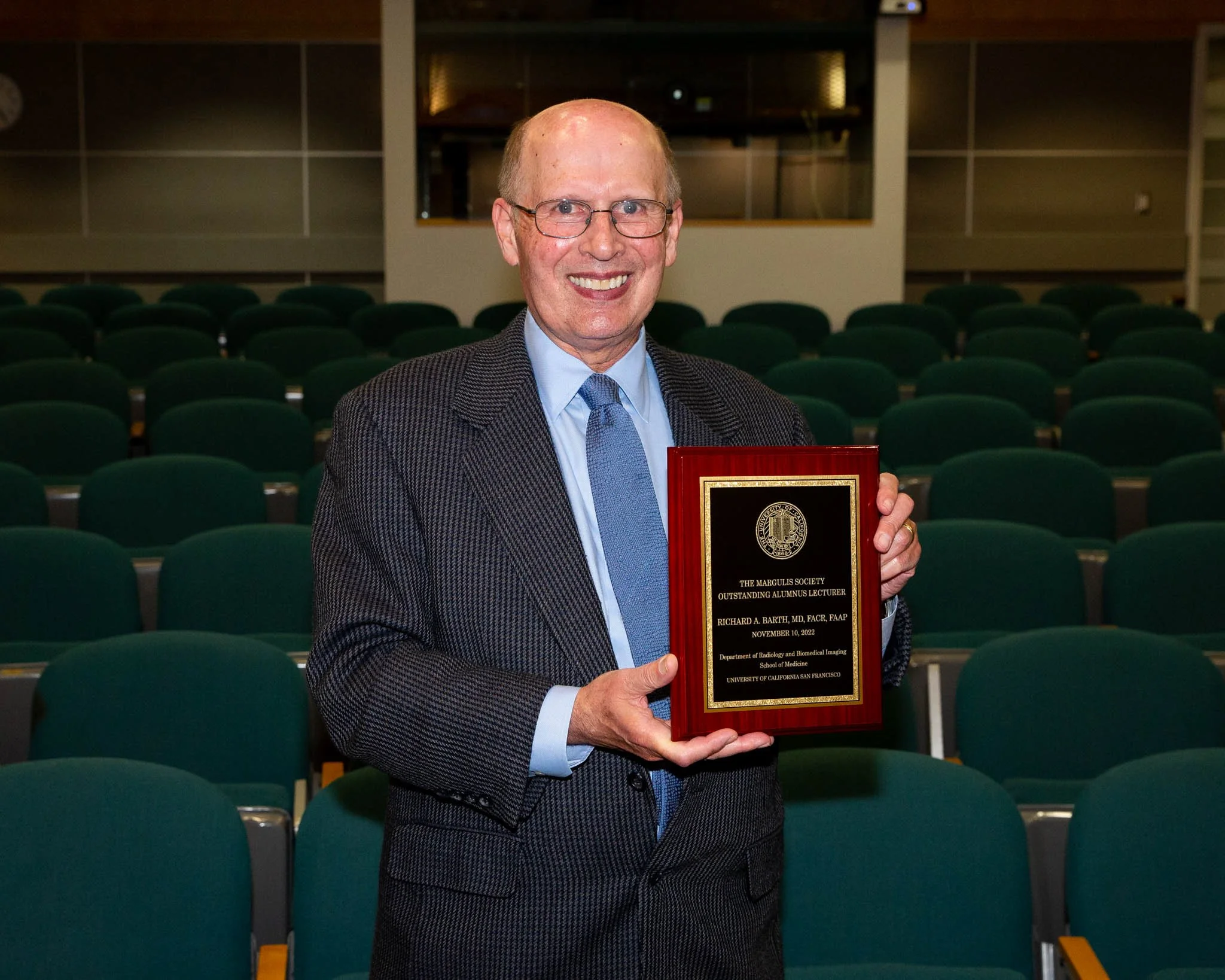 A smiling man in a suit holding a plaque in an auditorium with green seats.