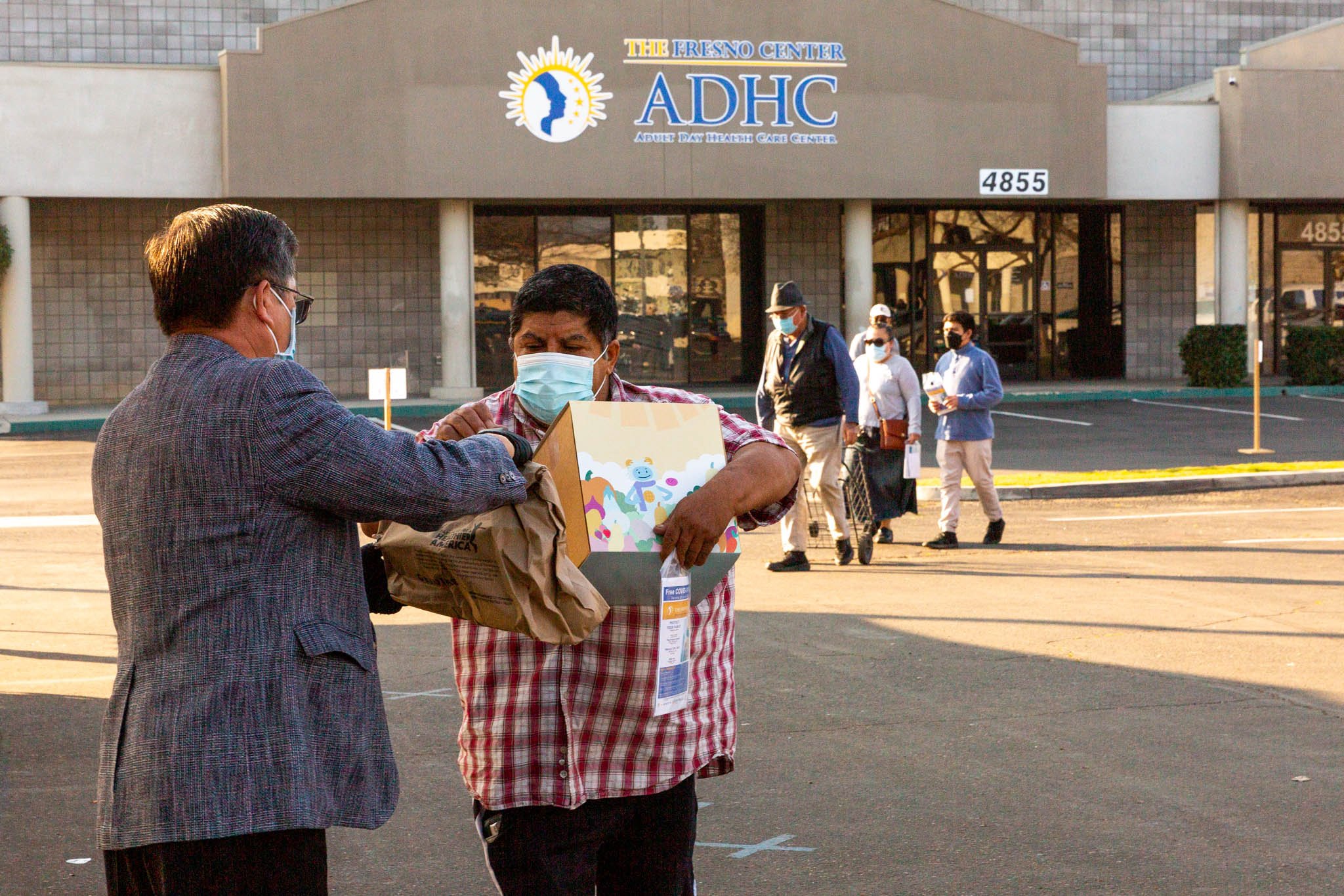 People wearing masks standing outside a building with a sign that reads 'The Fresno Center ADHC' and an address 4855, with some people walking in the background.