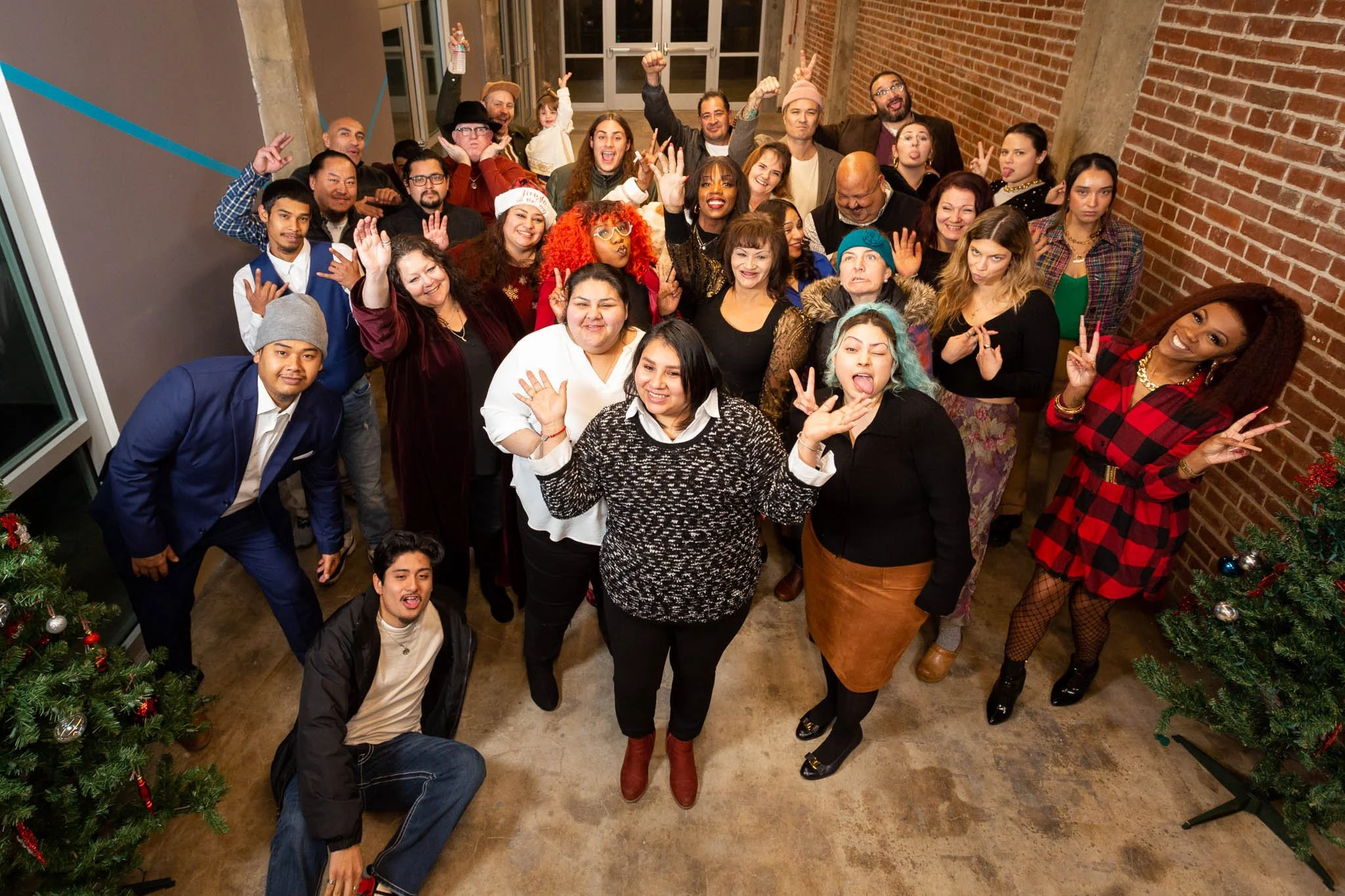 A large group of diverse people celebrating together in a festive setting with Christmas trees, smiling, making peace signs, and waving at the camera in a decorated indoor space.