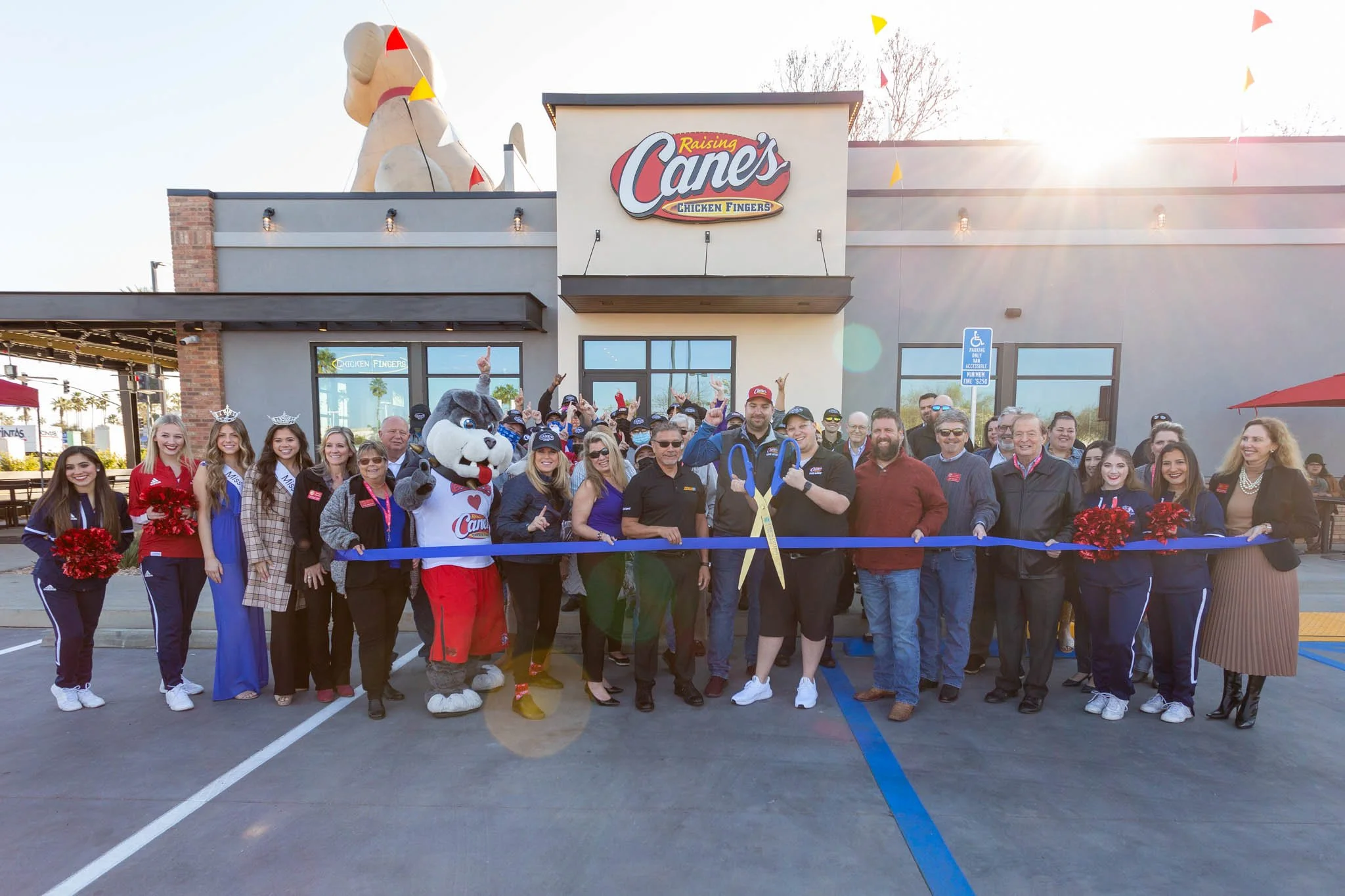 Group of people standing in front of Raising Cane's restaurant during a ribbon-cutting event, with a mascot, some wearing sashes and crowns, and cheerleaders holding pom-poms.