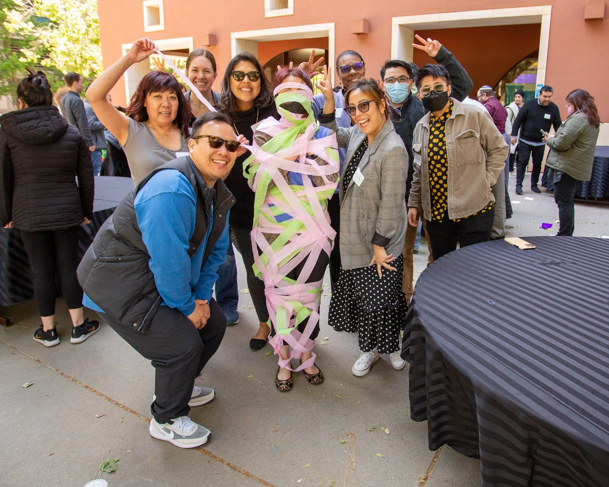 A group of people smiling and posing around a person wrapped in colorful ribbons, outdoors at a social gathering or party.