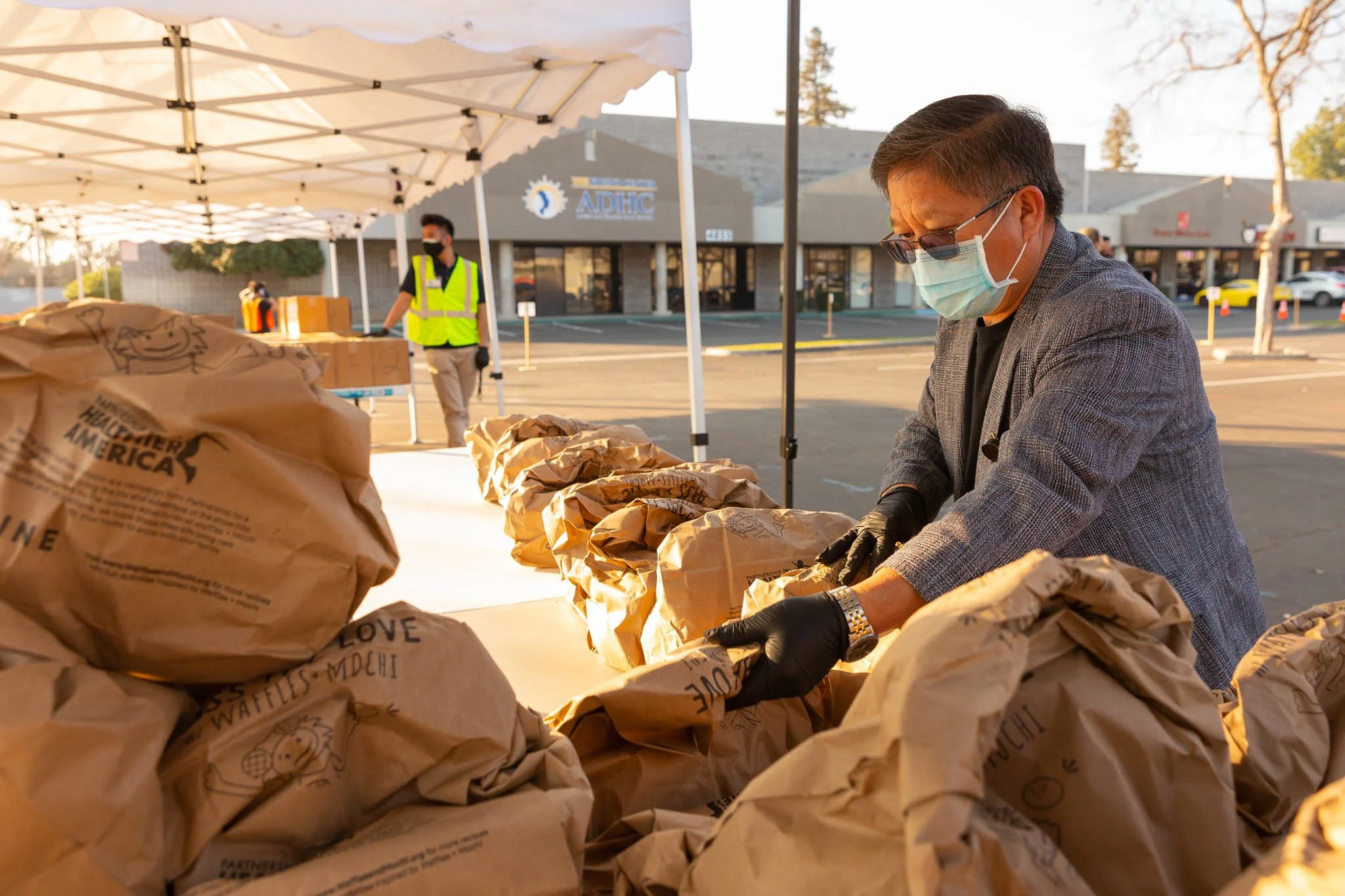 A man in a gray blazer, black gloves, face mask, and glasses organizes brown paper bags at an outdoor event, with a tent and two other people in the background, in a parking lot.