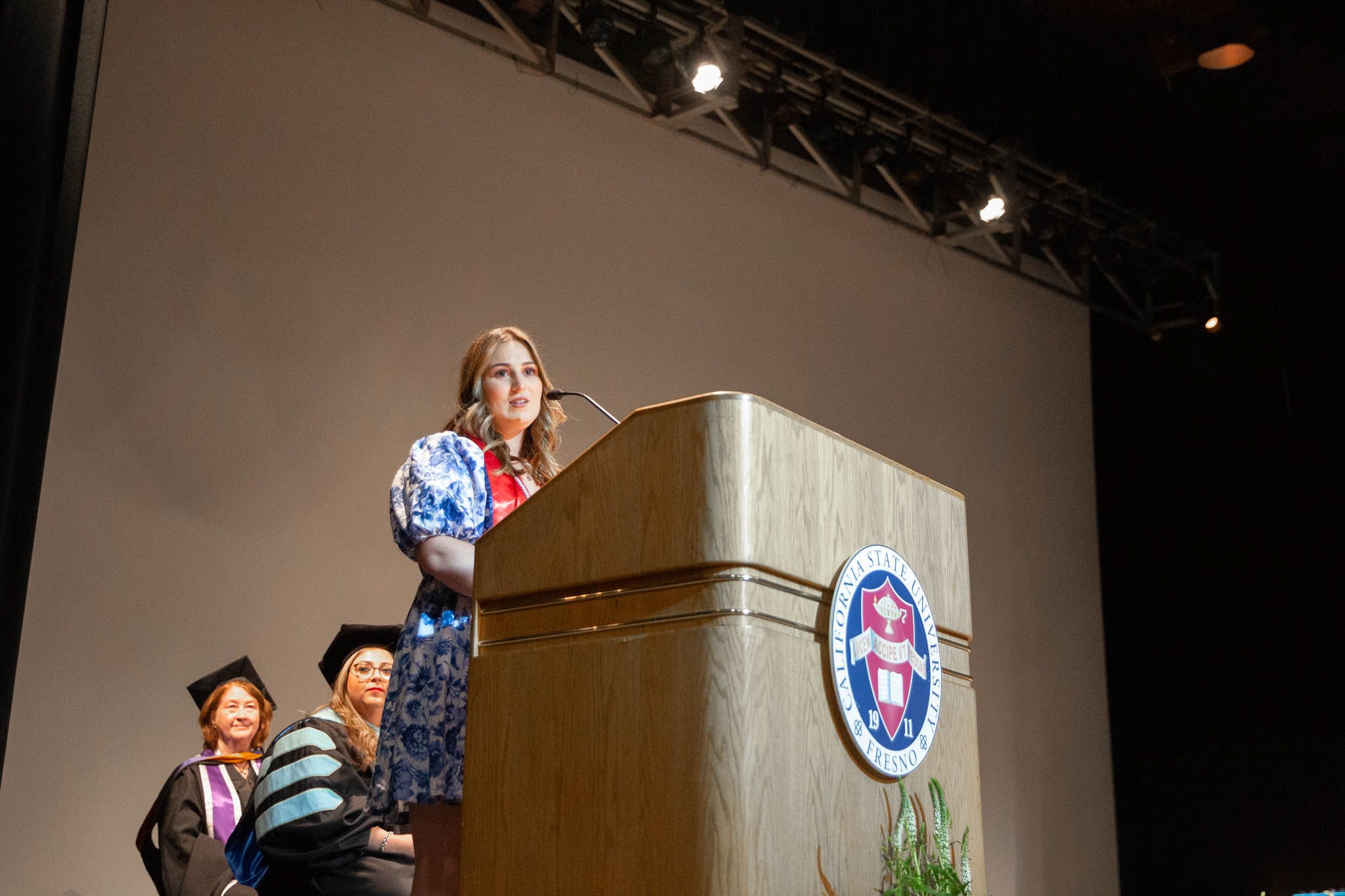 A young woman in a floral dress speaking at a university graduation ceremony, standing behind a wooden podium with the Fresno State seal, with three women in graduation gowns seated behind her.