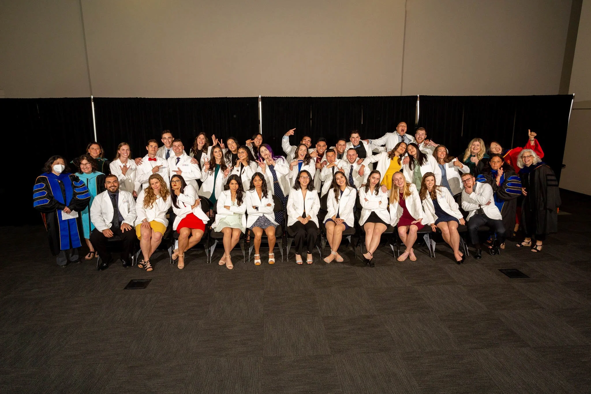 Group of graduates in white coats, some with academic gowns, posing for a photo on stage at a graduation ceremony.