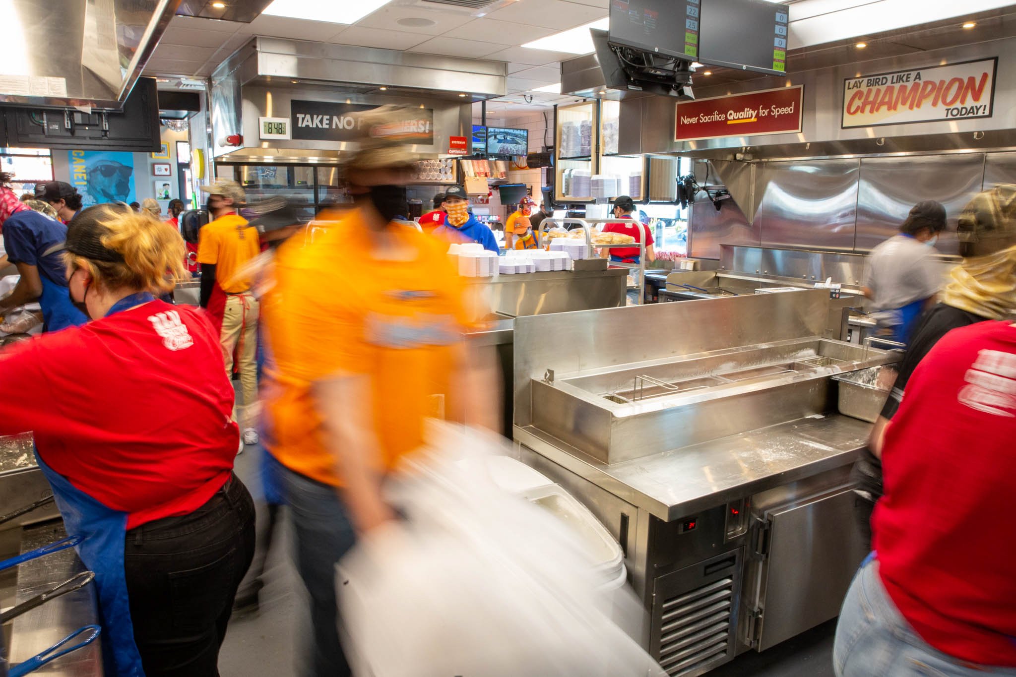 Inside a busy fast-food restaurant kitchen with workers preparing food. Some workers wear red, yellow, and blue uniforms with caps, and some are wearing masks. There are stainless steel appliances, a monitor displaying orders, and signs on the walls.