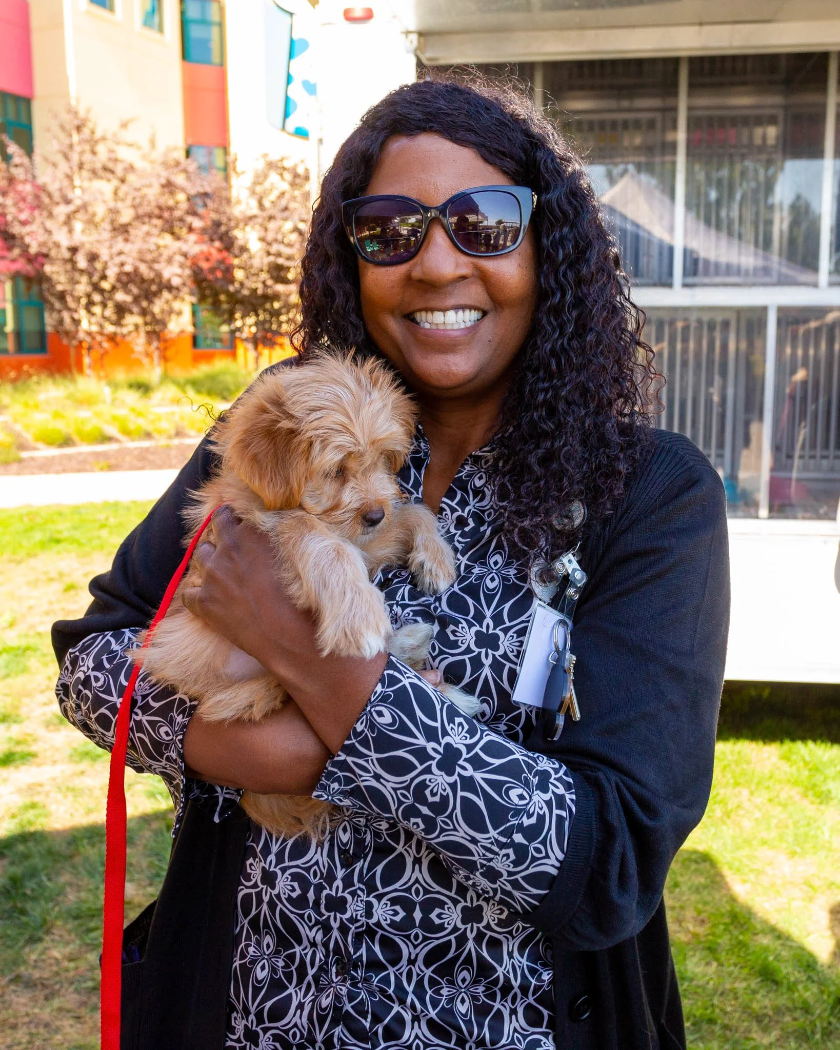 A woman with curly hair wearing sunglasses holding a puppy outside on a sunny day.