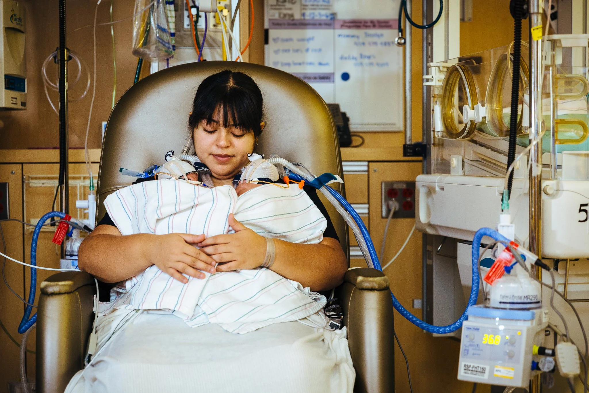 A woman sitting in a hospital chair holding a small patient, surrounded by medical equipment and monitoring devices.
