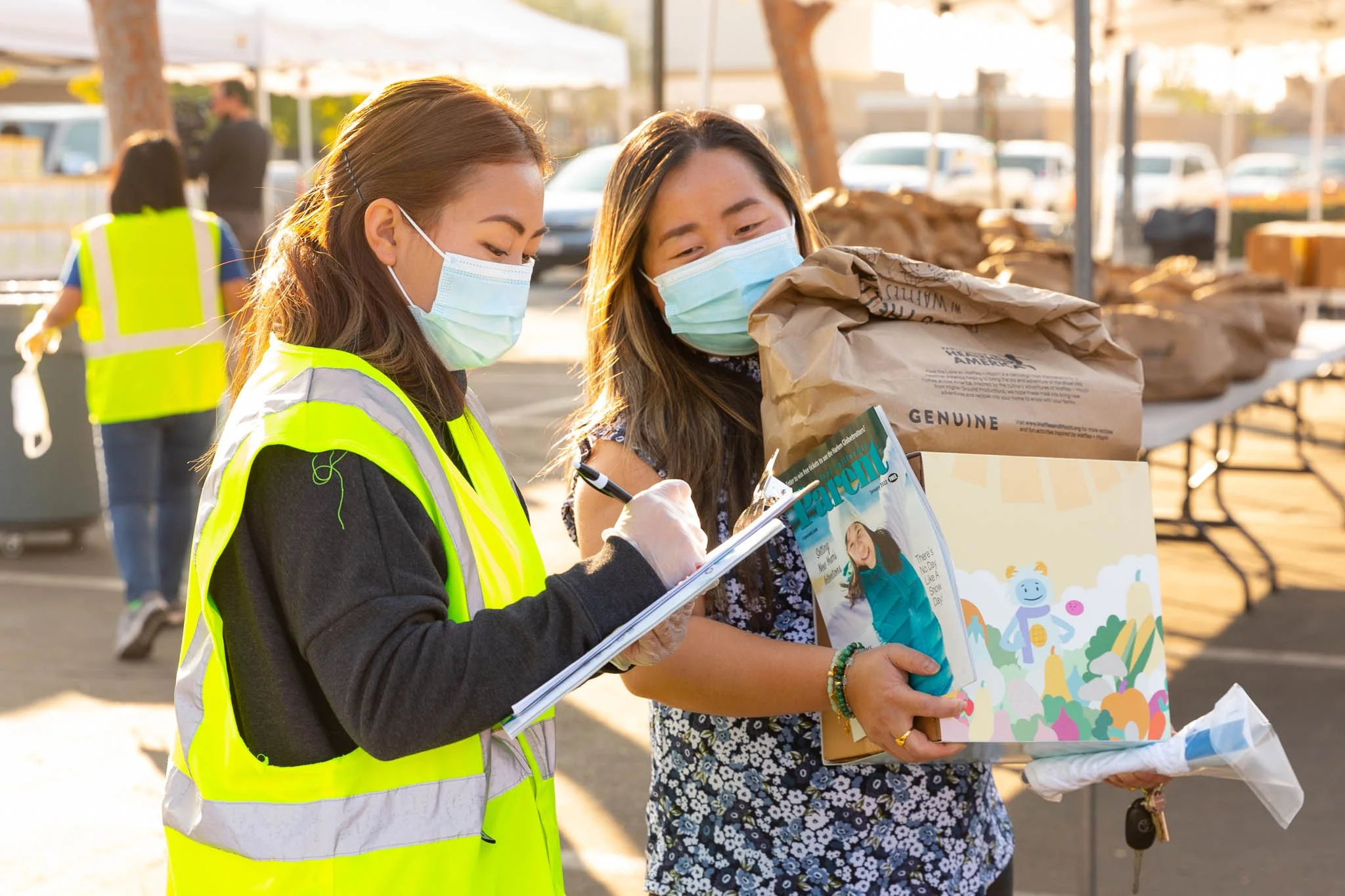 Two women wearing masks and yellow safety vests inspecting and writing on a clipboard at a food distribution event.