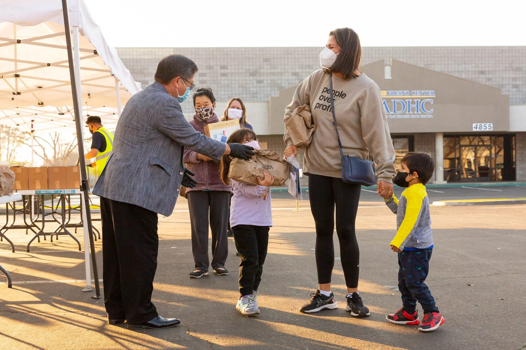 A group of people, including children and adults, are outdoors at a charity event. They are wearing masks, and one person is handing a bag to a woman holding a child's hand. There are tables and tents set up nearby.