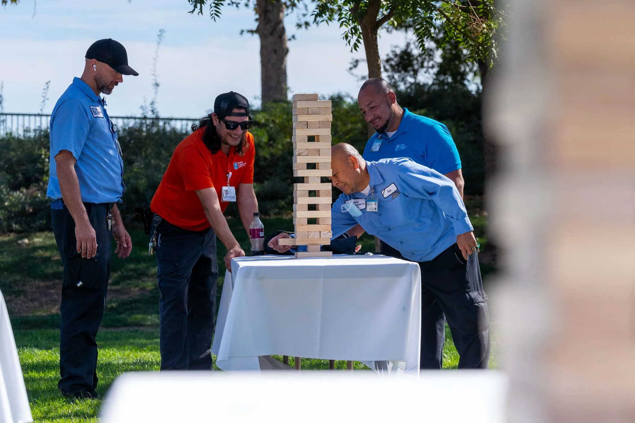 Four men playing giant Jenga outdoors at a table with a white tablecloth, with trees and blue sky in the background.