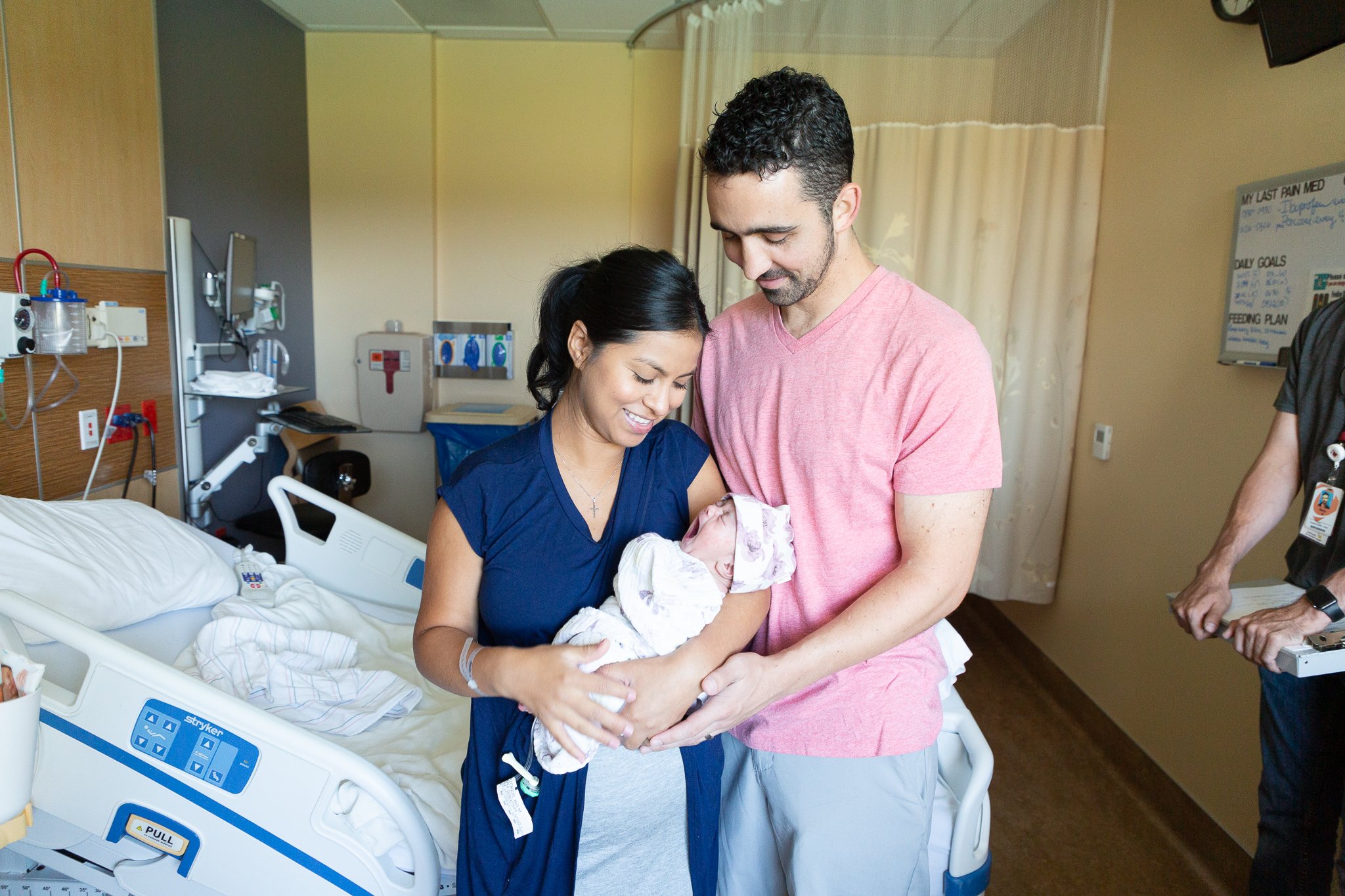 A woman in a hospital bed holding a newborn baby, with a man standing beside her and smiling at the baby. The hospital room has medical equipment and a whiteboard in the background.