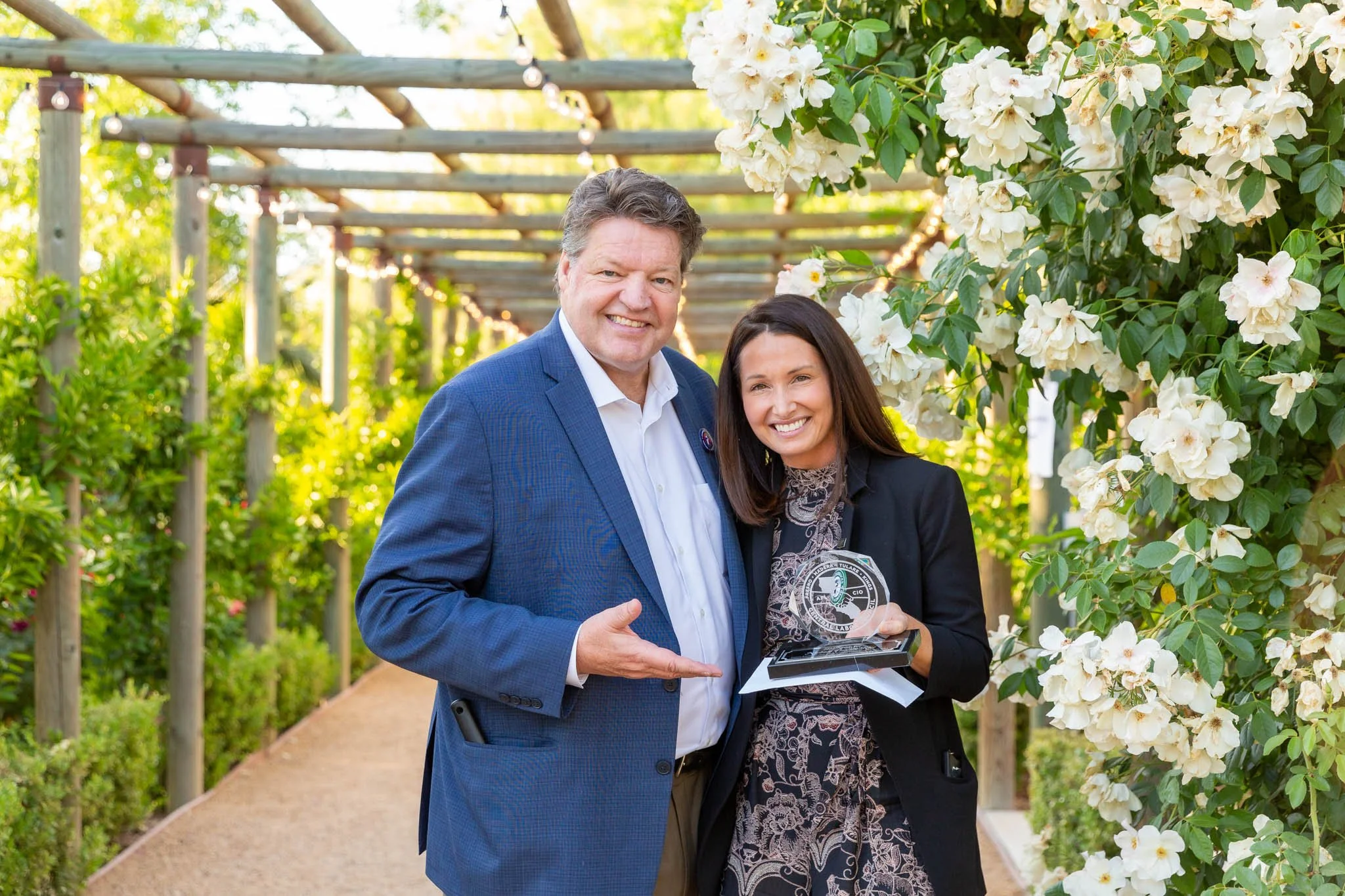 A man and a woman standing together outdoors in a flower garden, smiling at the camera, with the woman holding an award or plaque.