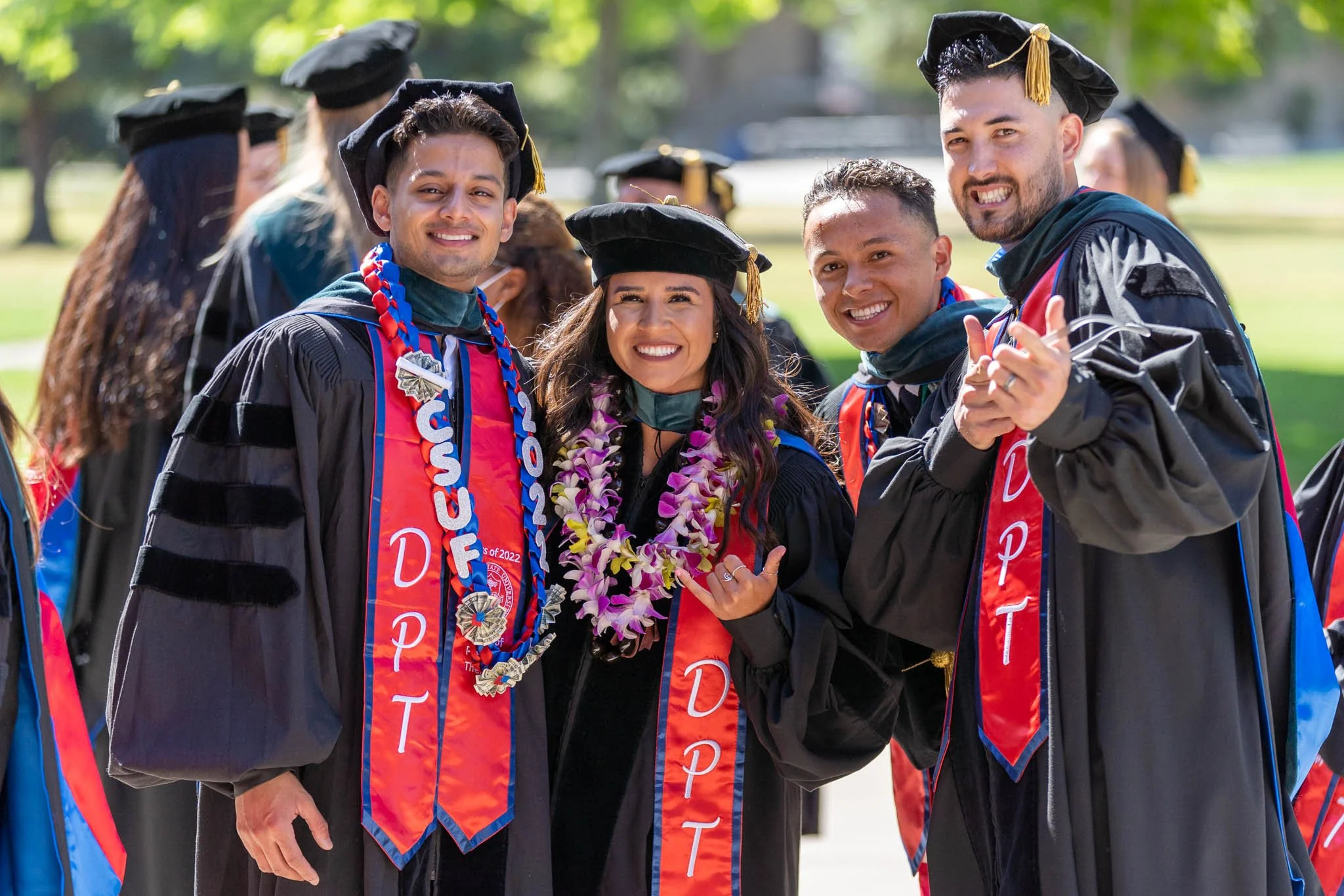Group of five diverse fresno state university graduates outdoors, dressed in caps and gowns, smiling, with some wearing honor cords and leis.
