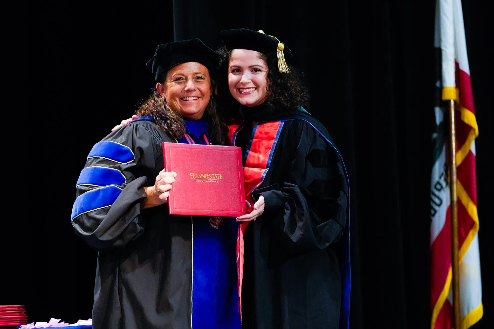 Two women in graduation gowns and caps smile and hold a red diploma cover that says 'Fresno State' on stage at a graduation ceremony, with a black backdrop and part of the California state flag visible.