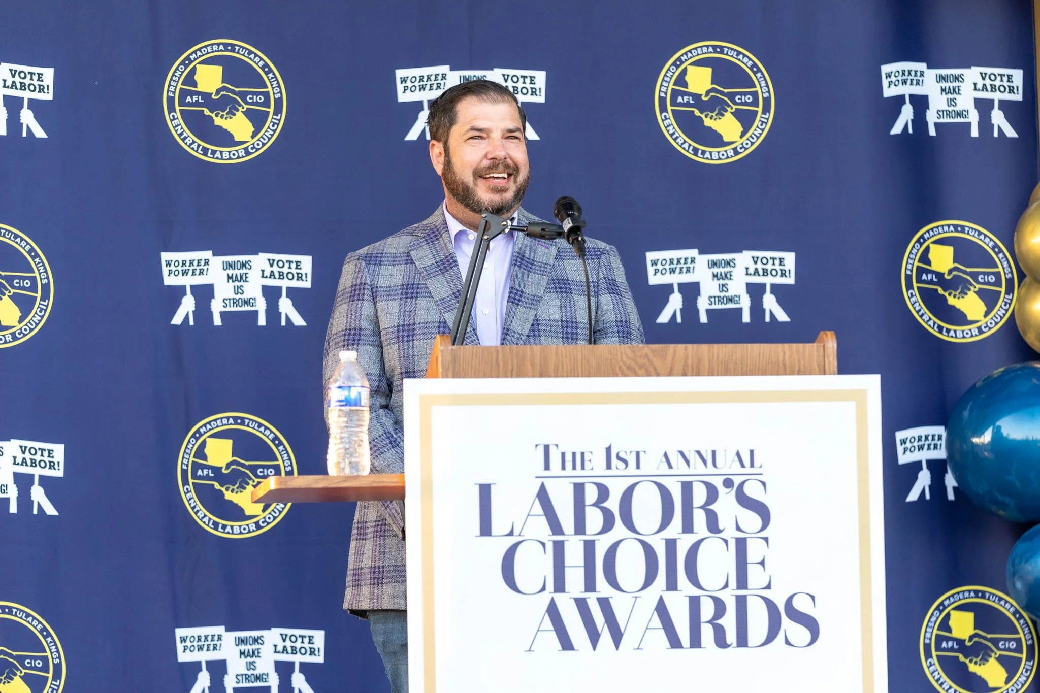 A man speaking at a podium during the 1st Annual Labor's Choice Awards. Behind him is a blue backdrop with logos of AFL-CIO and signs that read 'Vote Labor!,' 'Worker Power!,' and 'Unions Make Us Strong!'