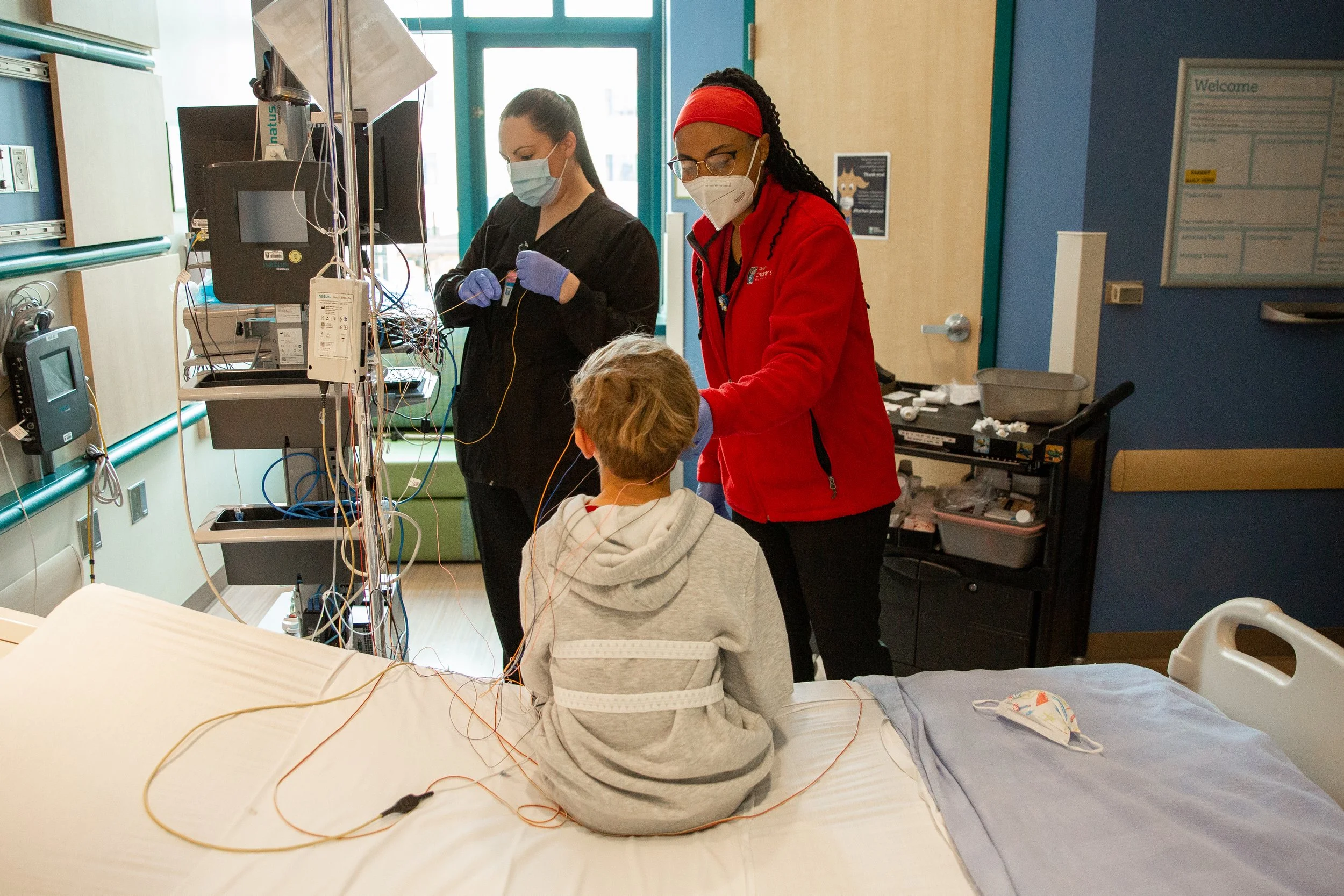 A young boy sitting in a hospital bed with medical wires attached, a nurse, and a medical technician in masks working with him.