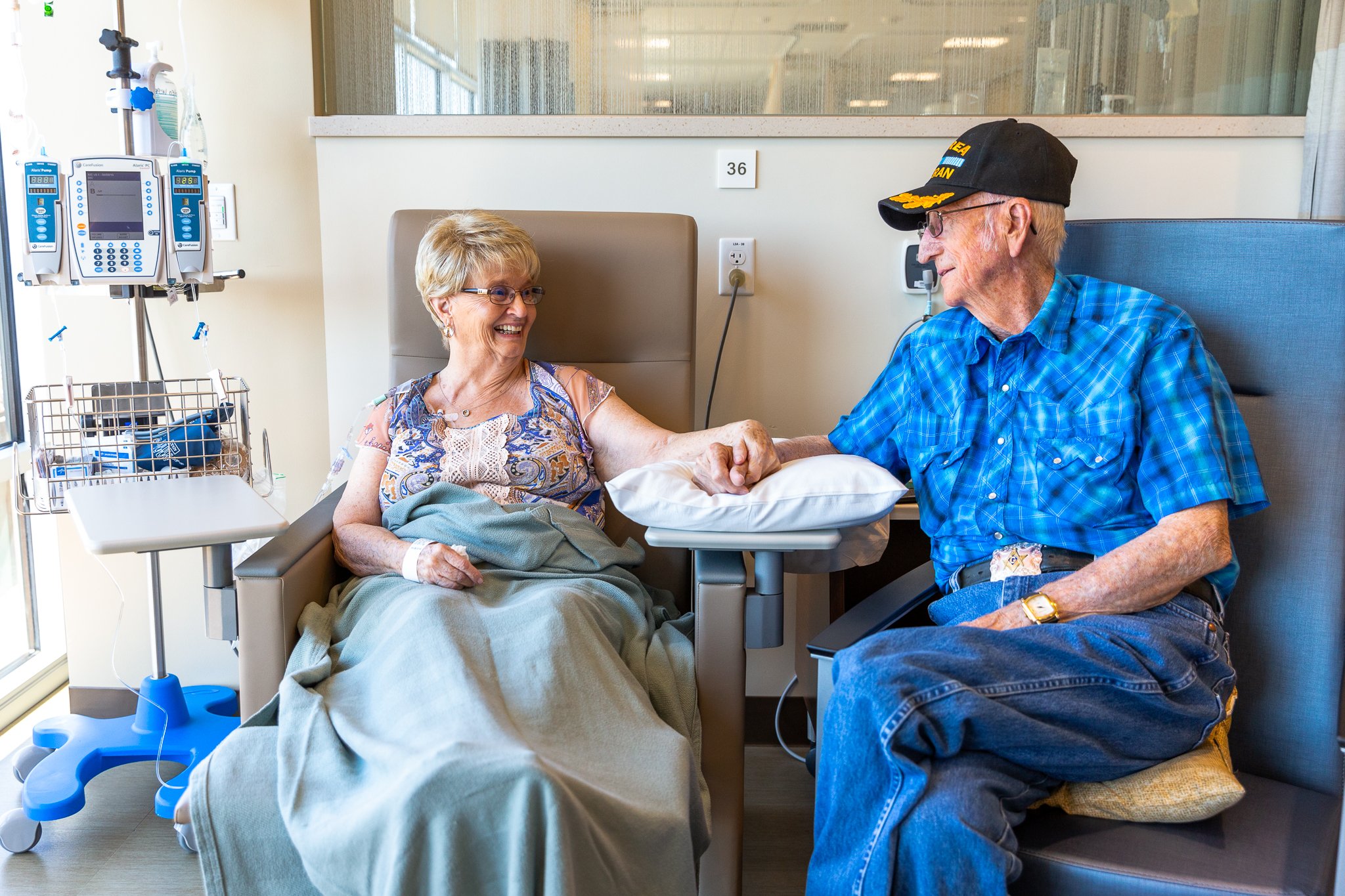 A woman and an elderly man sitting together in a hospital room, smiling and holding hands, with medical equipment nearby.