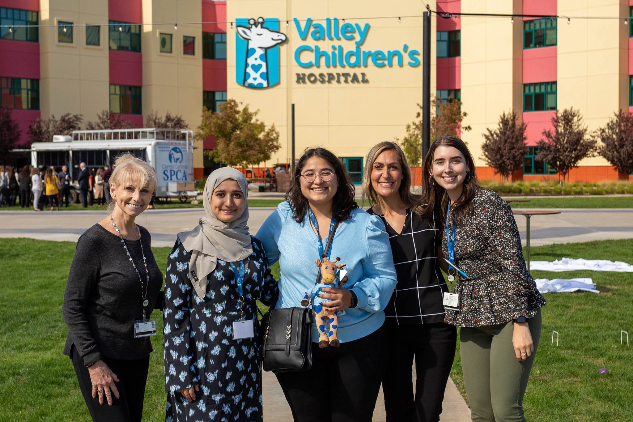 Five women standing outside Valley Children's Hospital, smiling for a group photo. They are dressed casually, with some wearing name tags. There are trees, a parked truck with SPCA branding, and a colorful hospital building in the background.