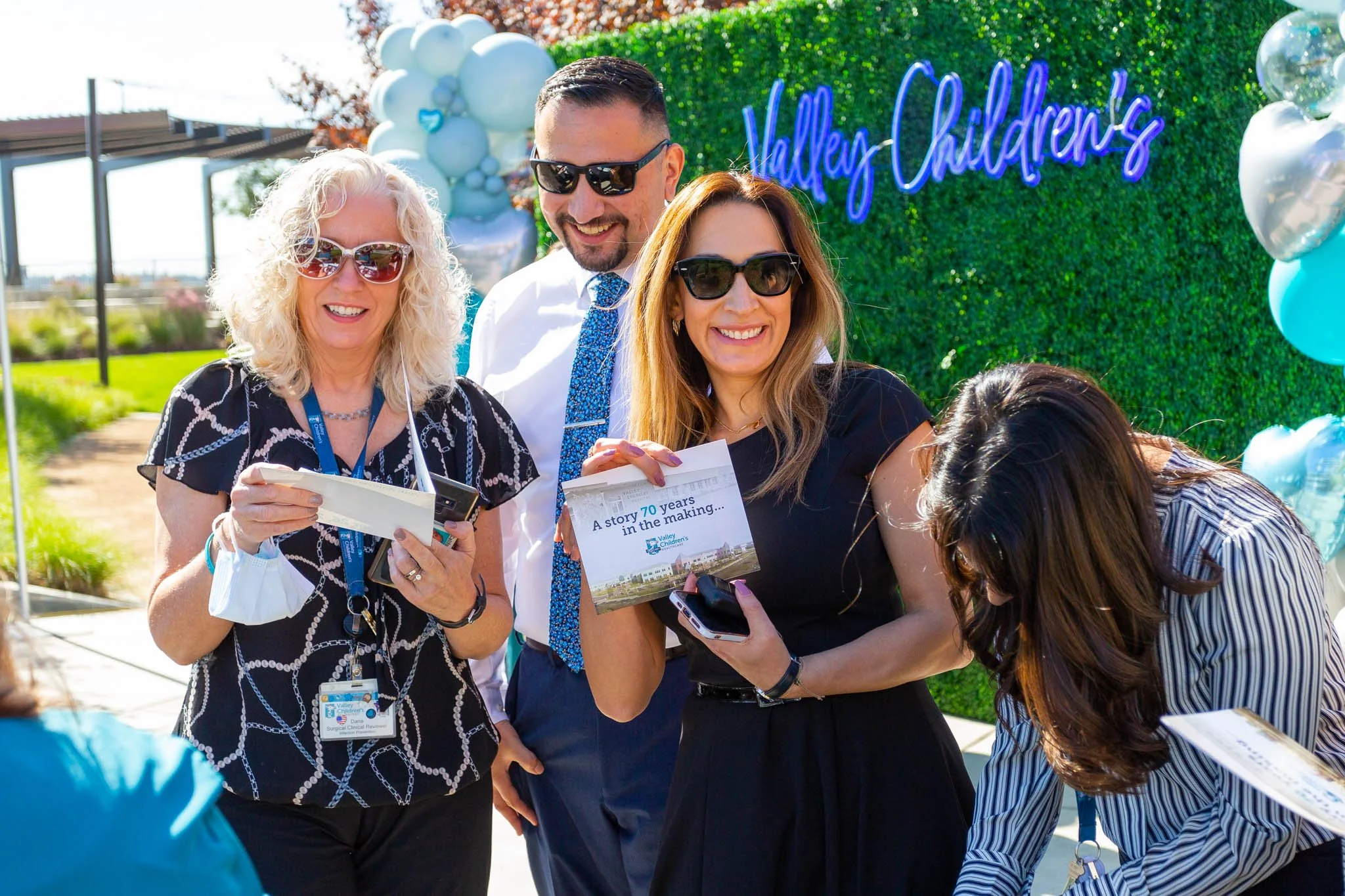 Group of five smiling adults, three women and two men, gathered outdoors during daytime at a celebration near a green hedge backdrop with a sign reading 'Valley Children's' and blue and silver balloons.