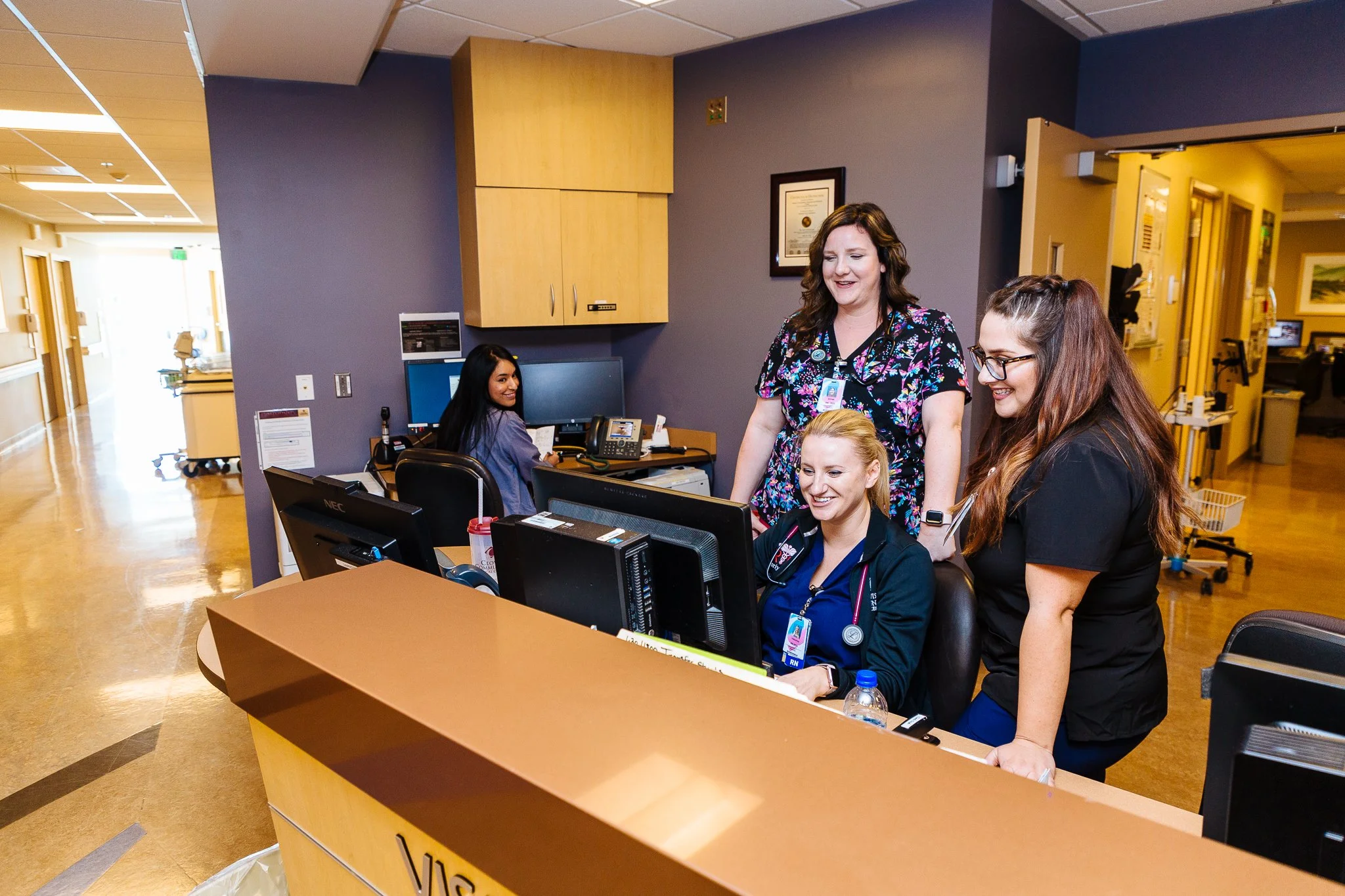 Group of four healthcare workers in a hospital reception area, with one seated at a computer and three standing around her, smiling and engaging with her.
